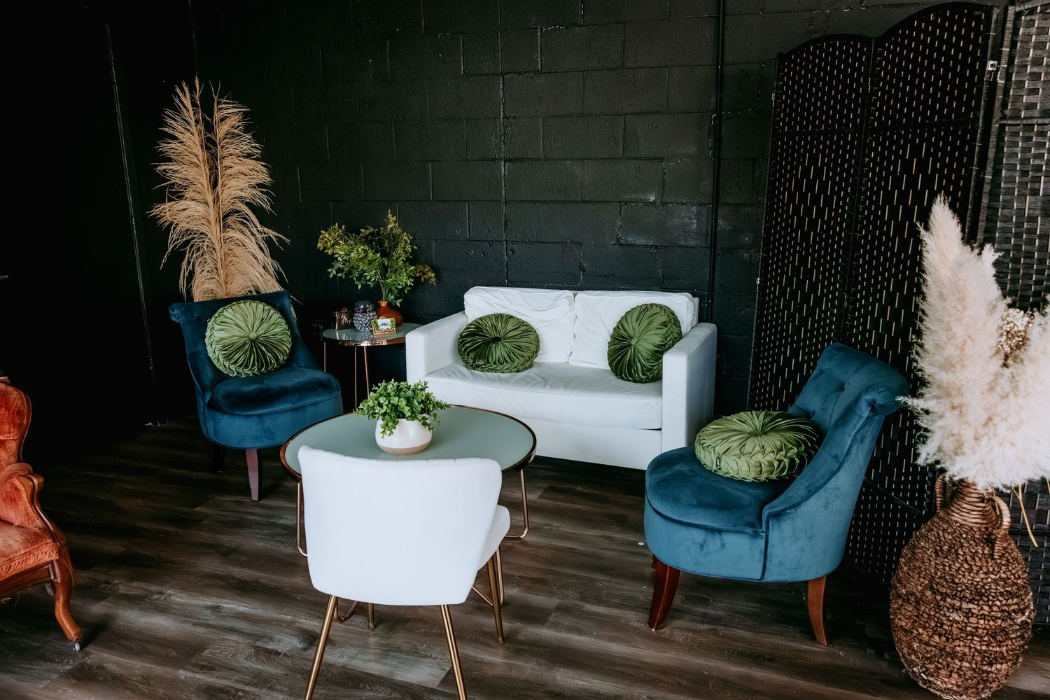 Elegant lounge seating area with white sofa, blue velvet chairs, green cushions, and pampas grass decor against dark walls.