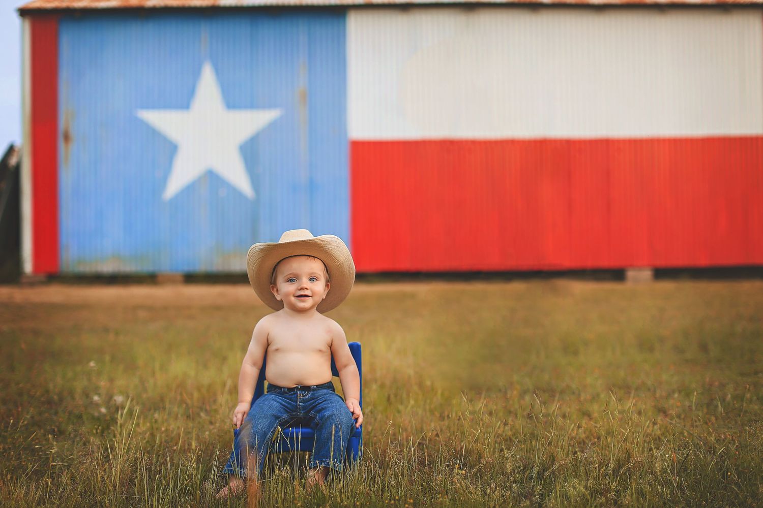 Shirtless toddler in cowboy hat and jeans sits in chair before a Texas flag painted barn.