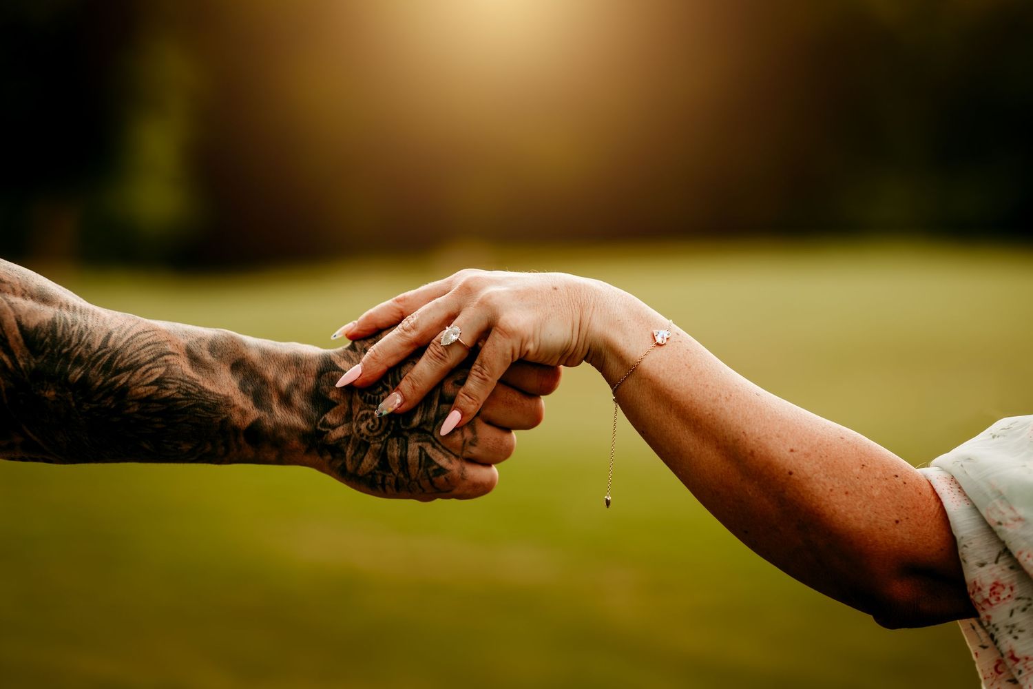 Two hands gently clasping, one with tattoos and one with a small wrist tattoo, set against a sunlit green background.