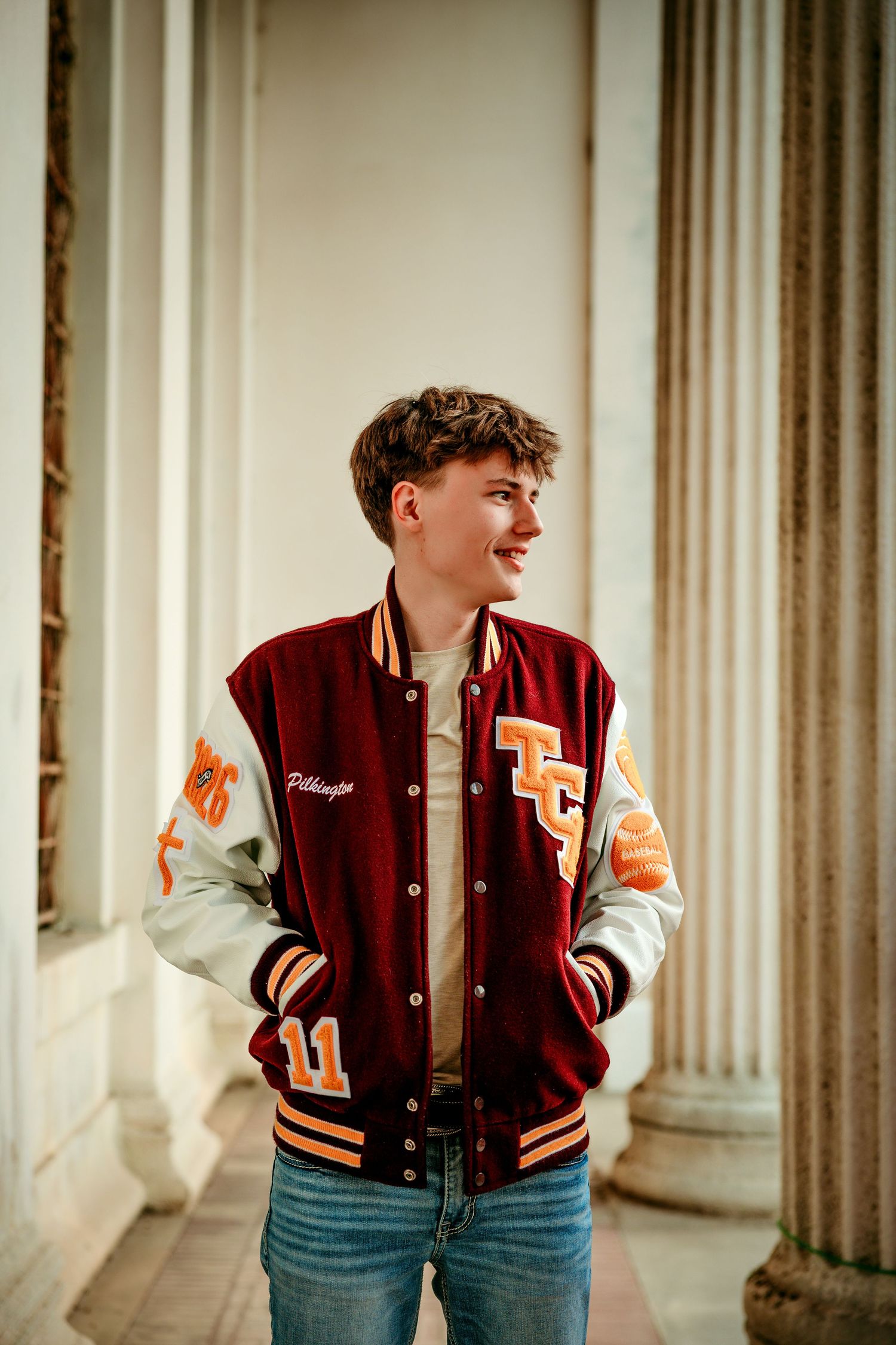 Young man smiling in a maroon varsity jacket with orange accents, standing between white columns.