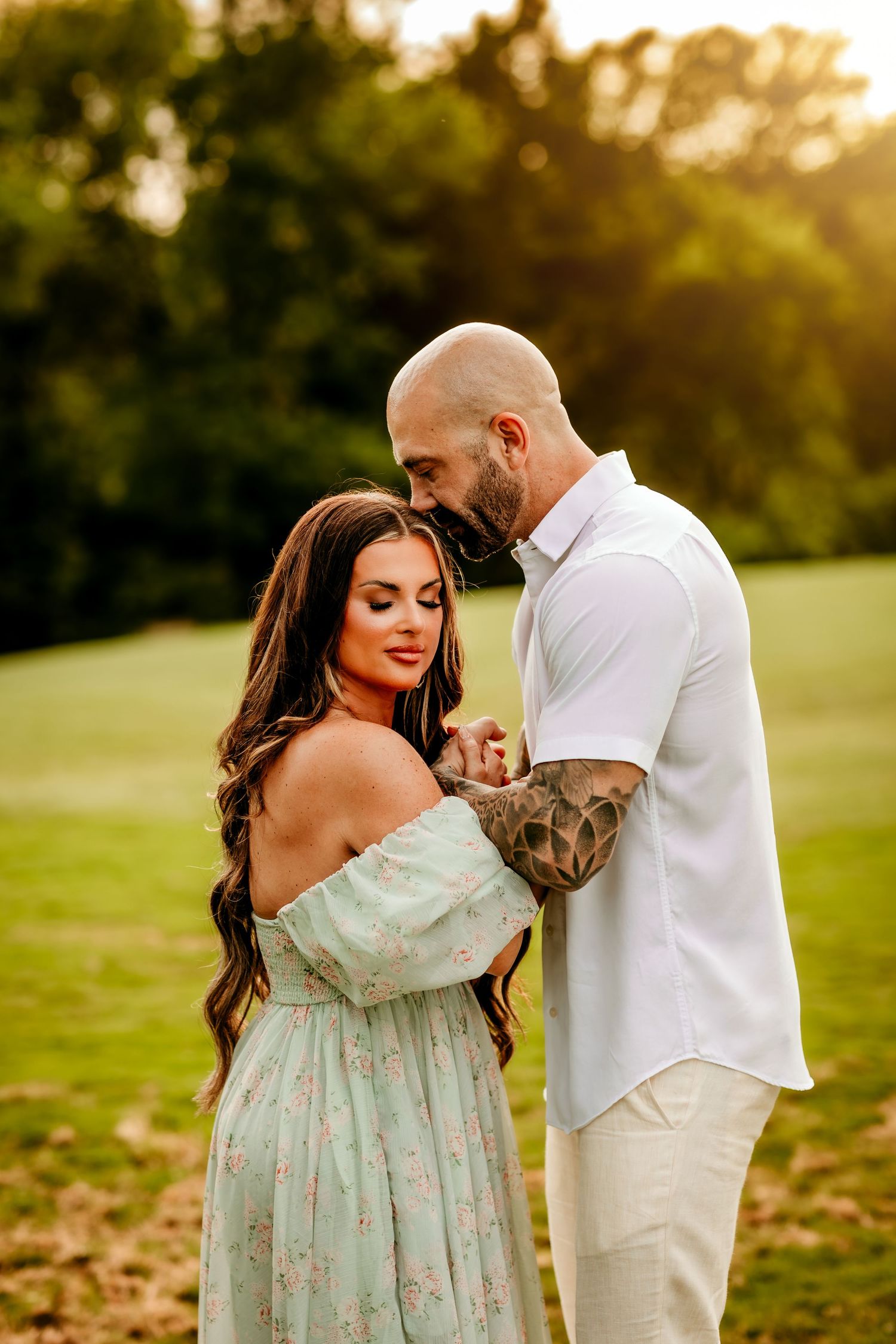 Couple sharing a tender moment at golden hour, woman in floral off-shoulder dress, tattooed man in white shirt.