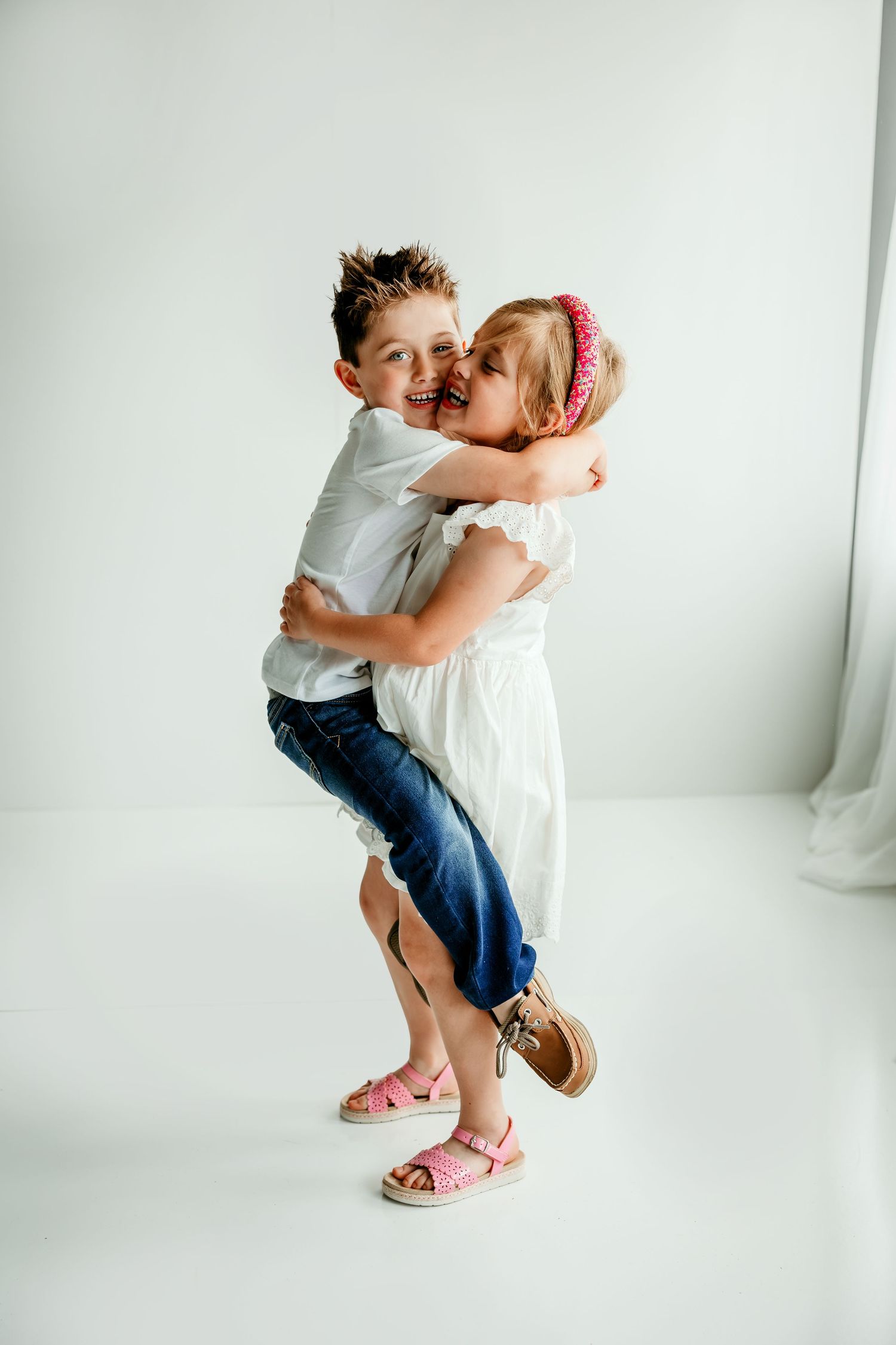 Two happy children hugging in a white studio, girl in white dress lifting smiling boy in jeans and white shirt.