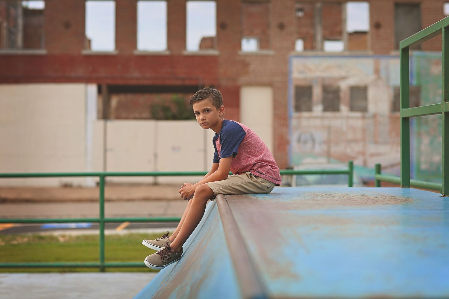 A young boy in a pink shirt and khaki shorts sits on a blue ramp at an outdoor skate park.