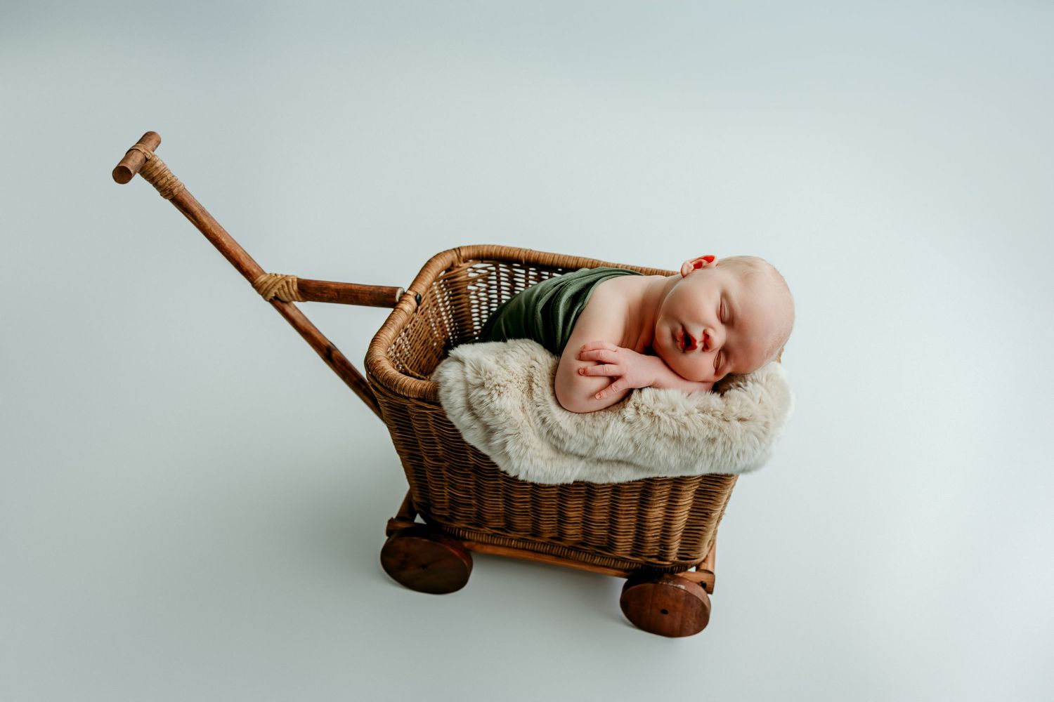 Sleeping newborn baby in green outfit nestled in a vintage wicker wagon with fluffy white blanket.