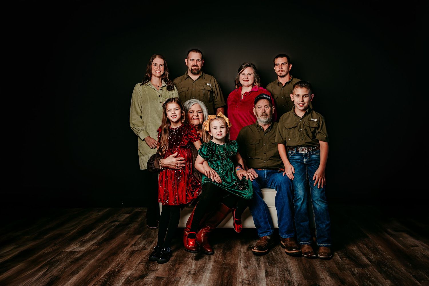 Family of eight poses for a festive holiday portrait in red and green outfits against a dark studio background.