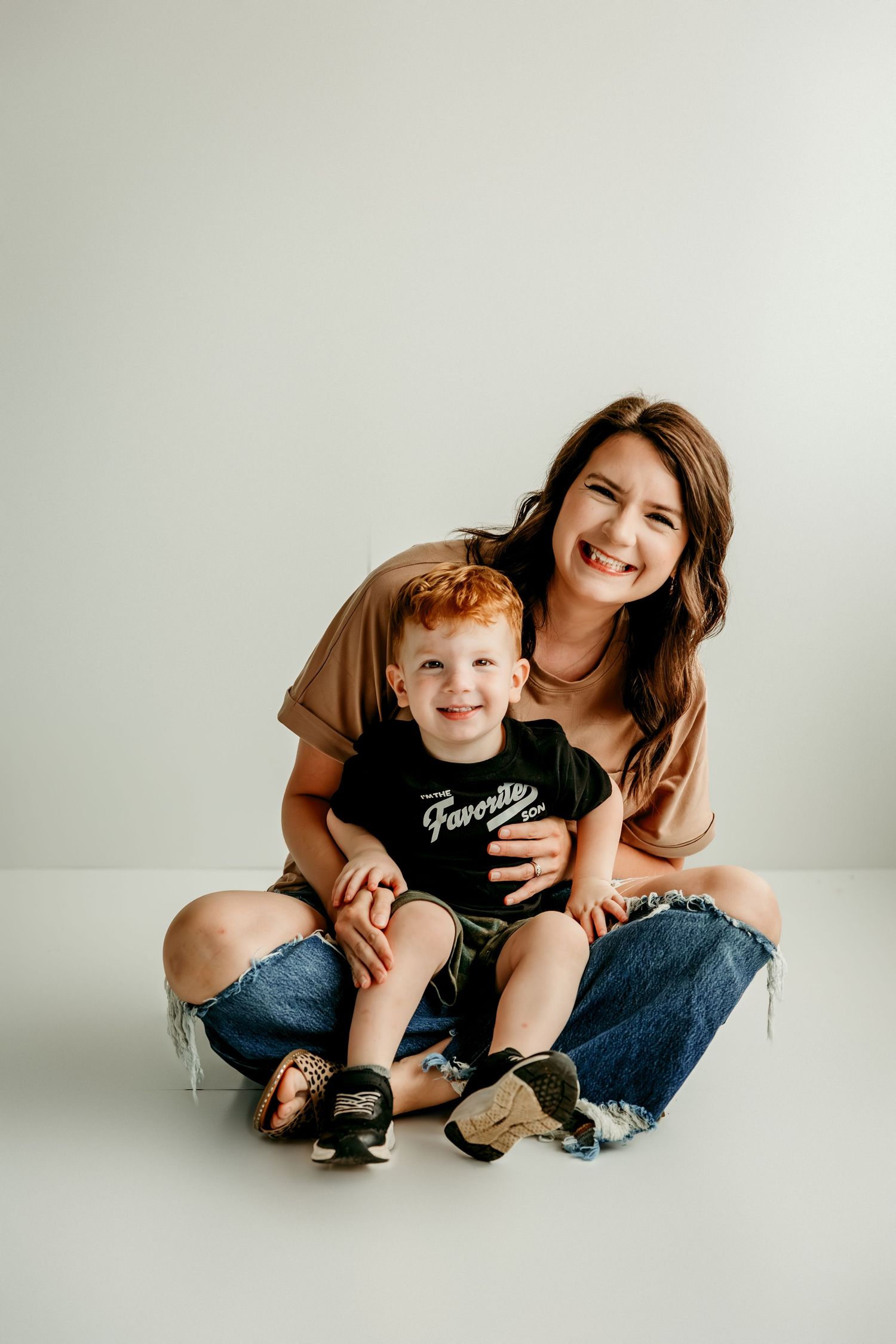 A smiling woman sits cross-legged holding a happy red-haired toddler boy in a studio portrait.