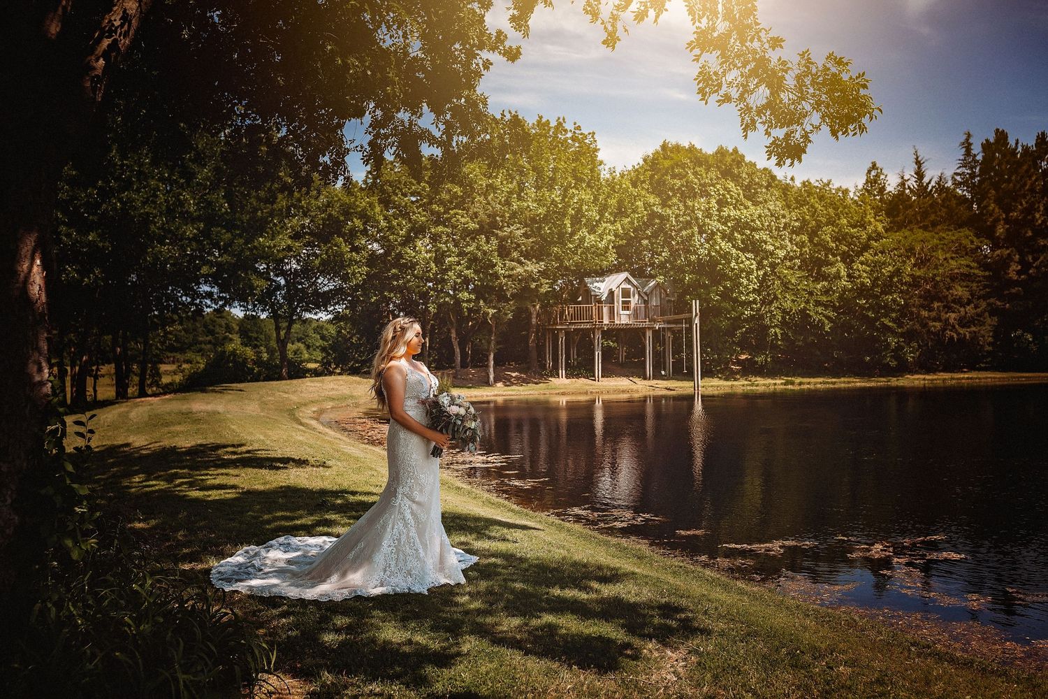 Bride in white gown stands by a sunlit pond with lush trees and a wooden structure in the background.