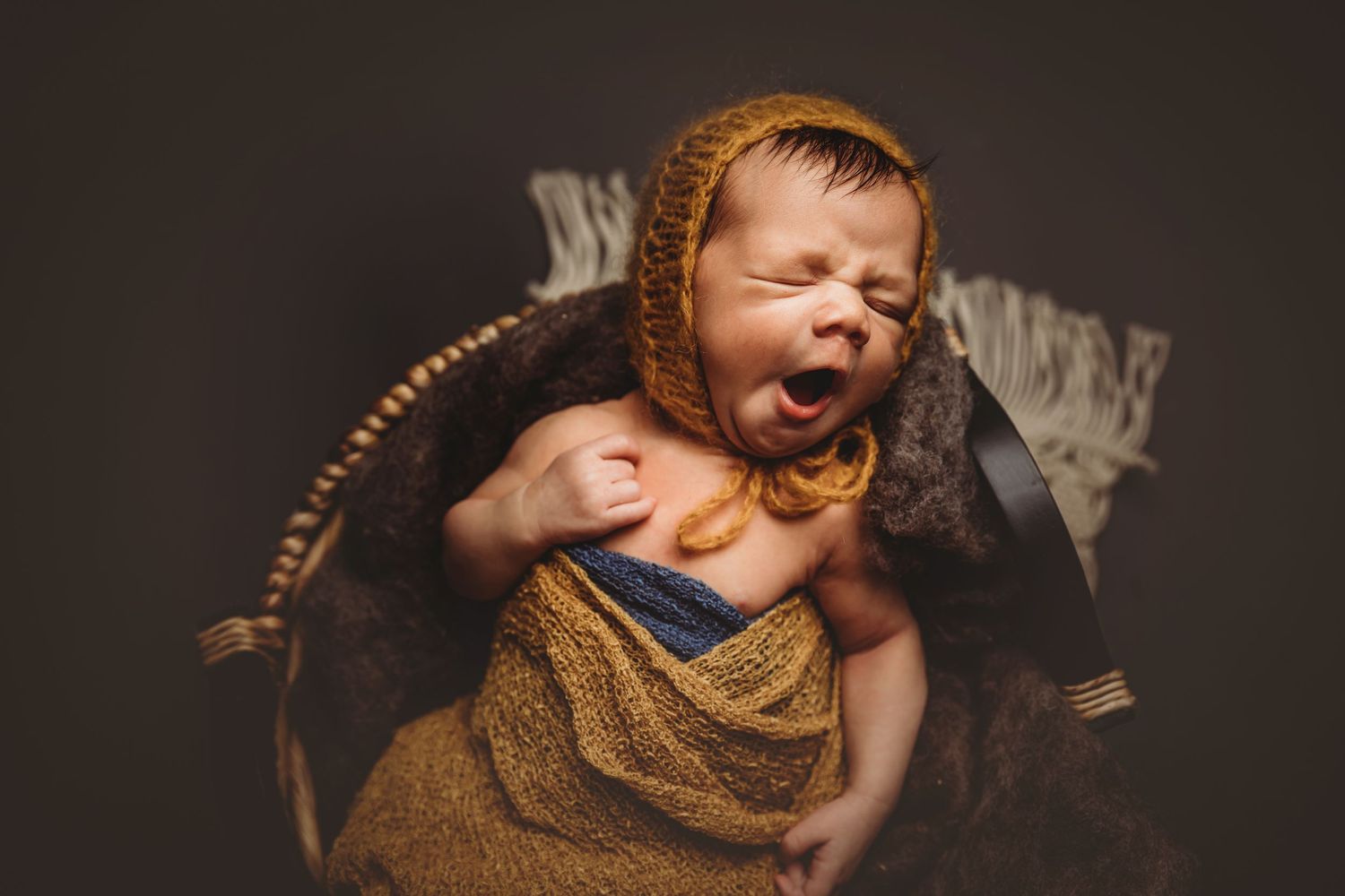 Newborn baby yawning in a wicker basket, wrapped in golden yellow fabric, wearing a knit bonnet.