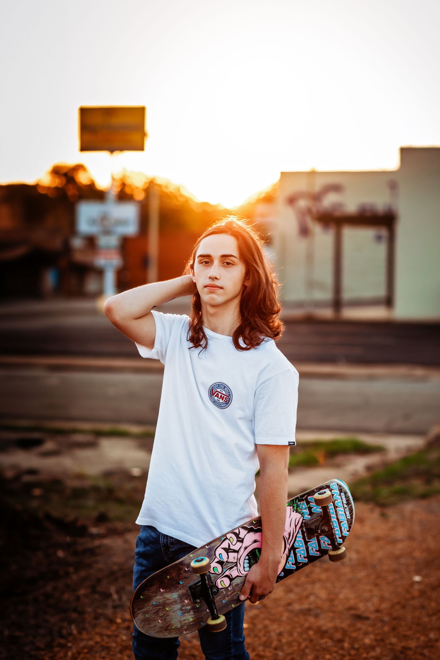 Young man with long hair holding a skateboard at golden hour sunset in an urban street setting.