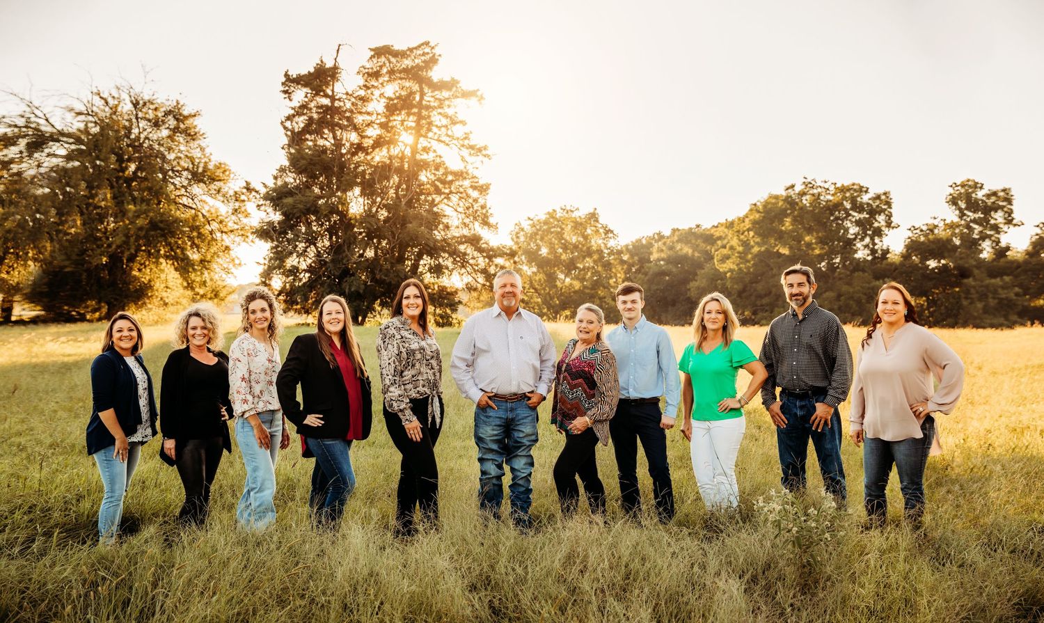 Group of ten people posing together in a golden sunlit field with trees in the background.