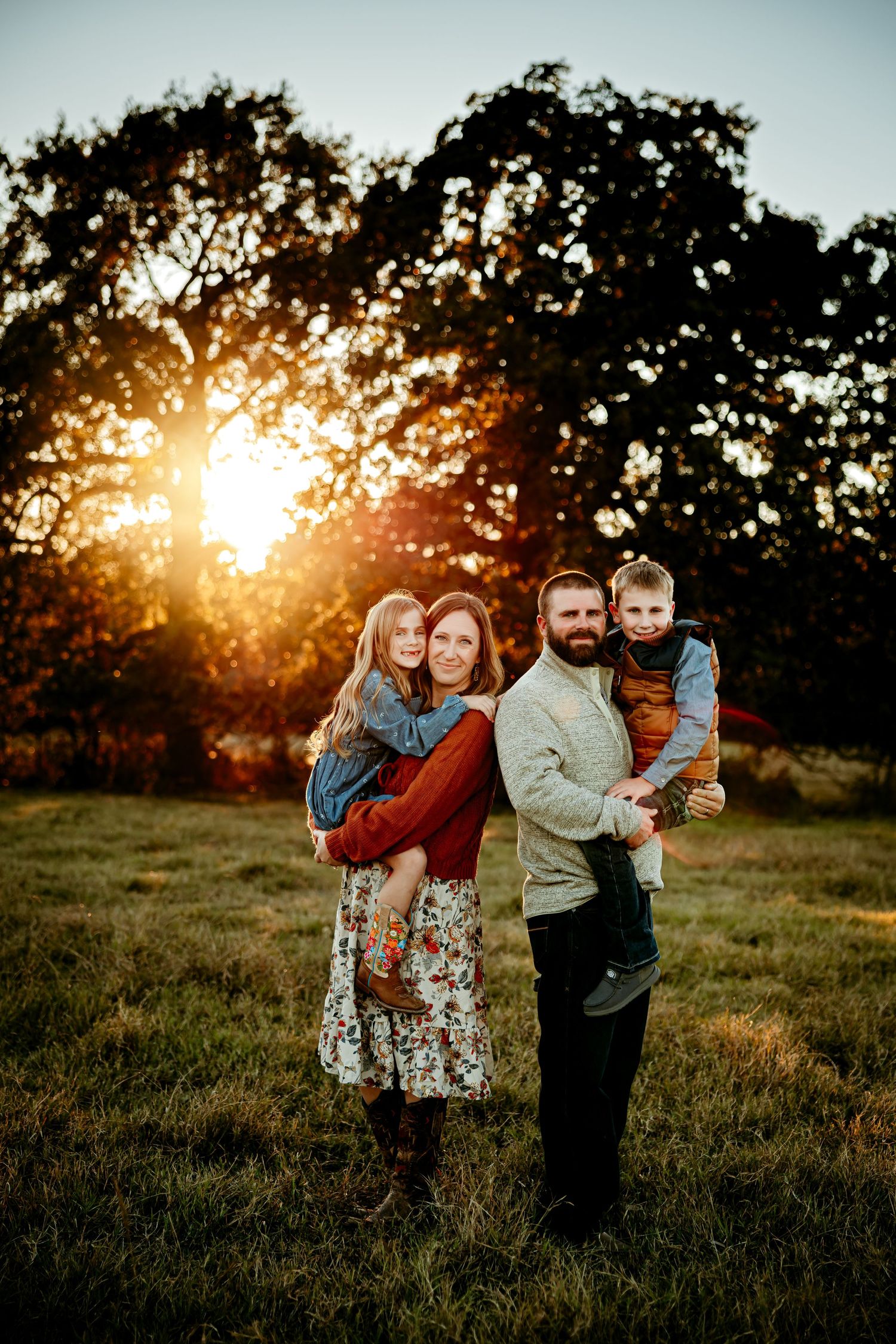 Family of four posing at golden hour sunset in a green field, parents holding young children, warm autumn tones.