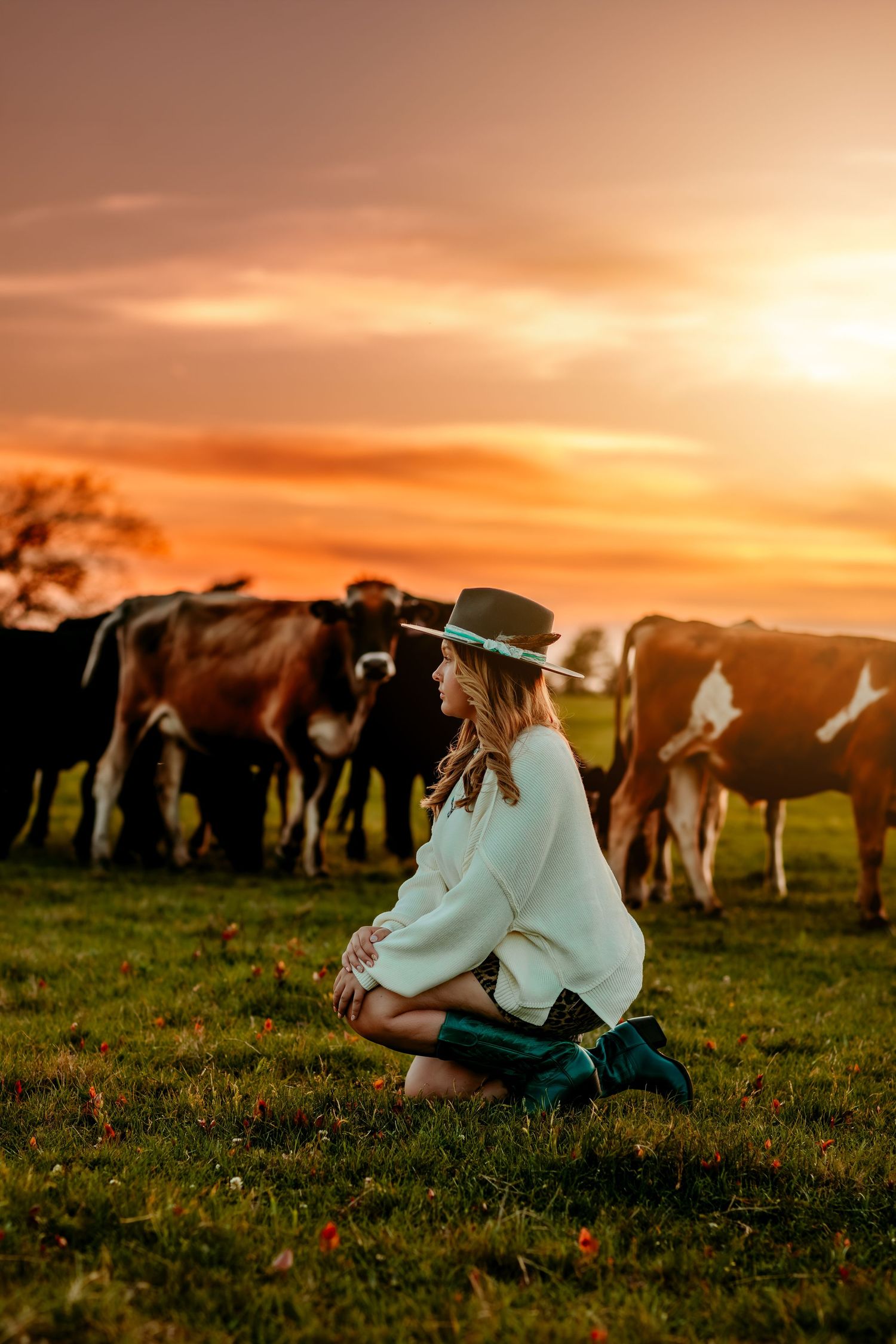 Woman in hat kneeling in green field at golden sunset with herd of cows grazing in background.