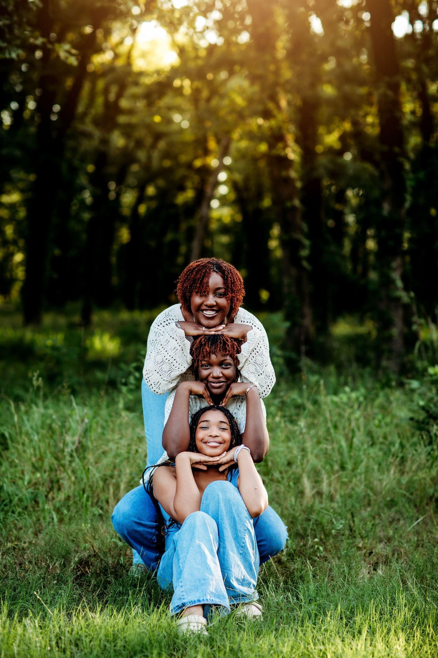 A joyful Black family of three poses outdoors in a sunny green forest, stacked playfully while smiling at the camera.