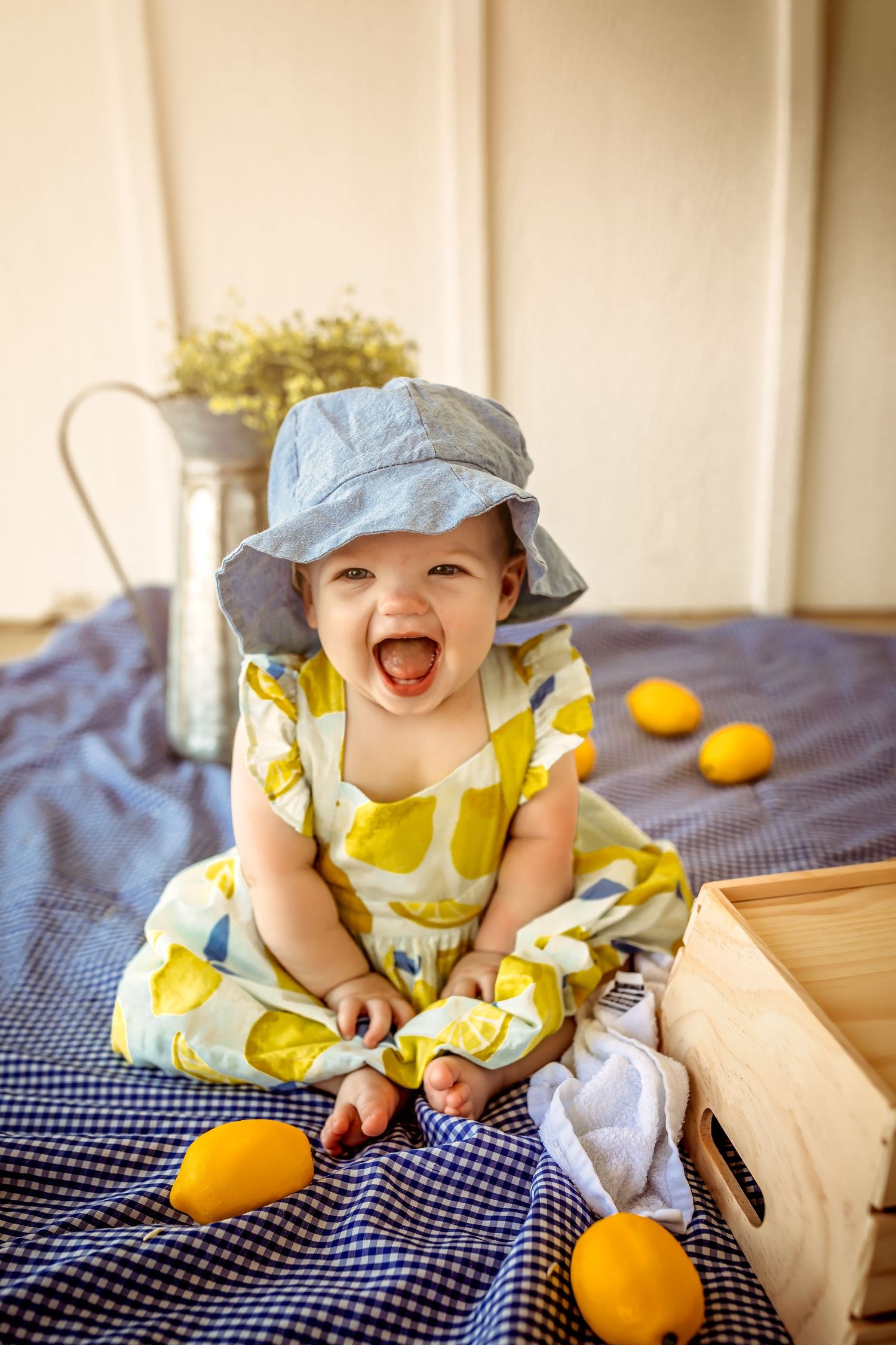 Happy baby girl in yellow lemon dress and blue hat sitting on blanket with fresh lemons, laughing joyfully.