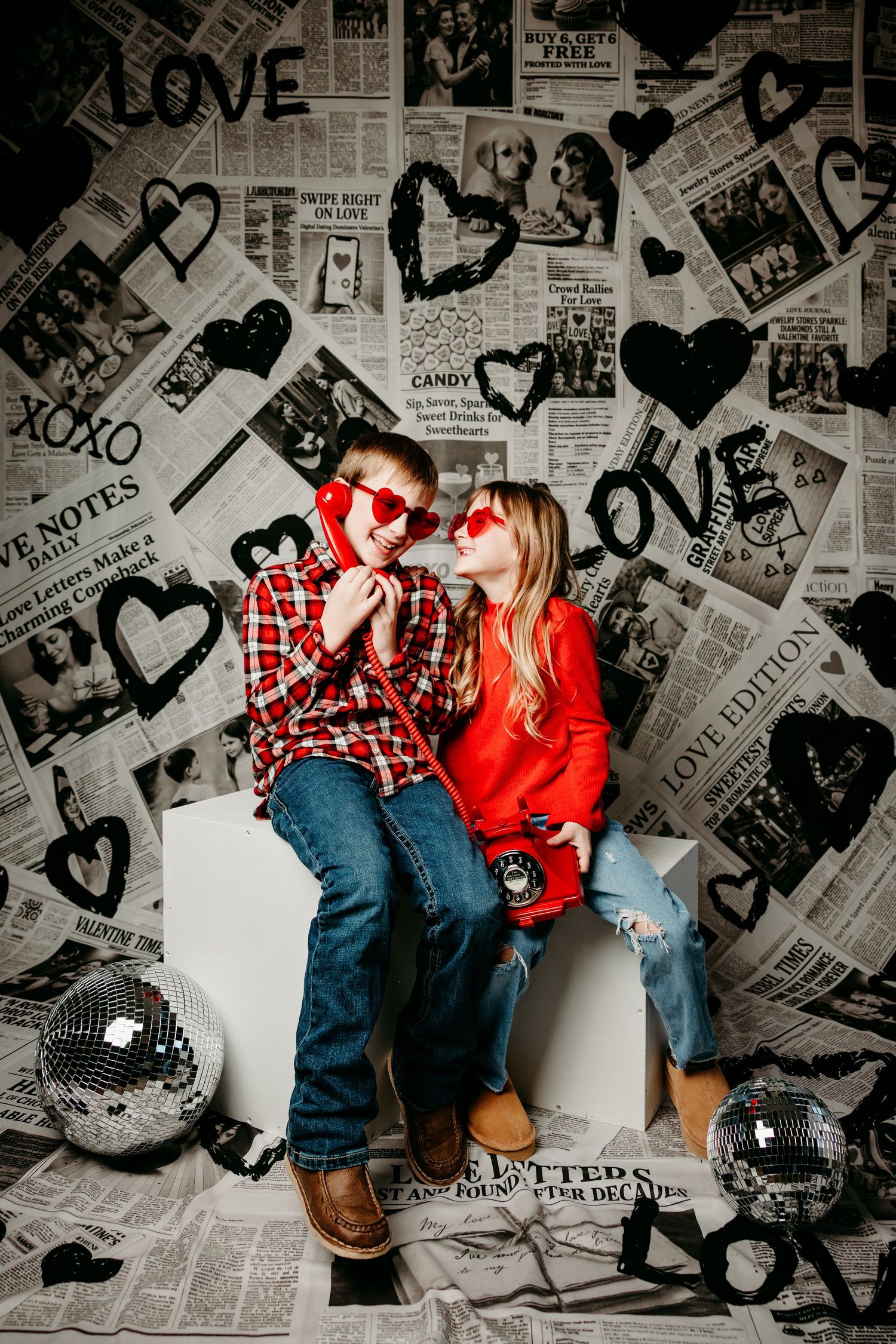 Couple in red and plaid outfits posing against newspaper wall with hearts in artistic black and white photoshoot series.