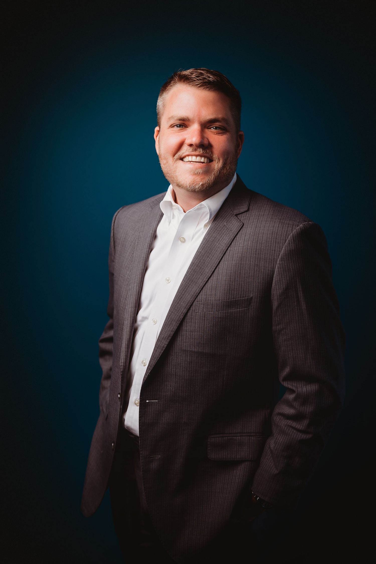 Smiling professional man in gray suit and white shirt poses confidently against dark teal studio background.
