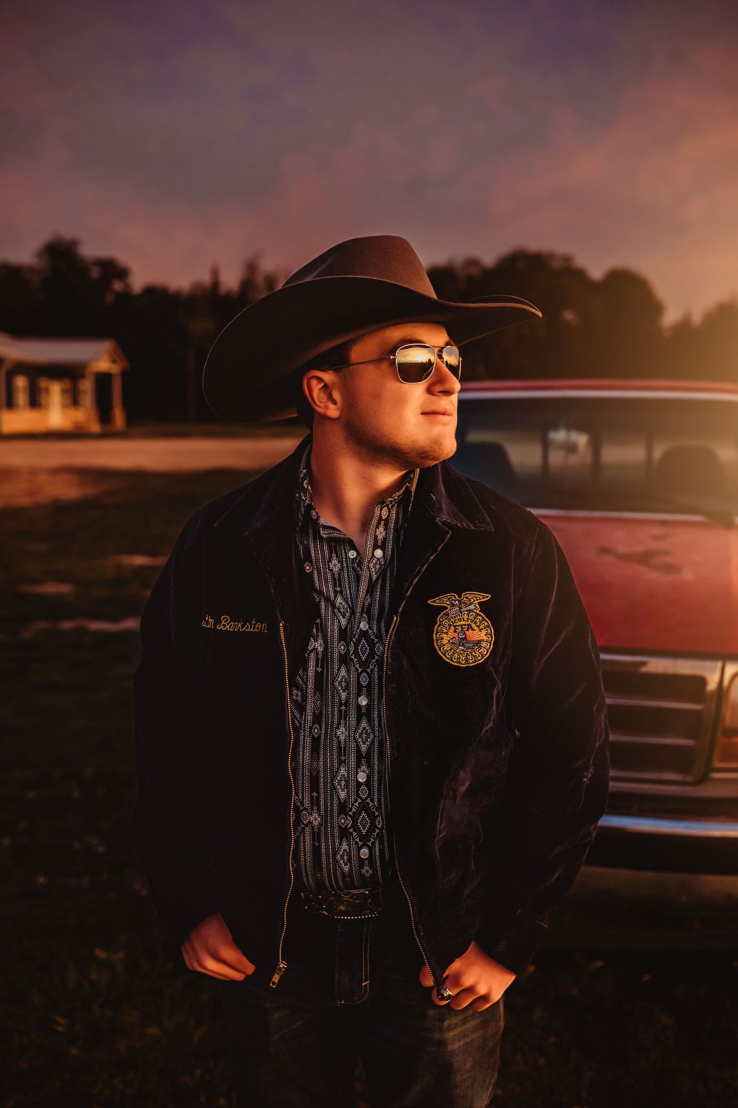 Young man in FFA jacket, cowboy hat, and sunglasses standing near a vintage truck at golden sunset.