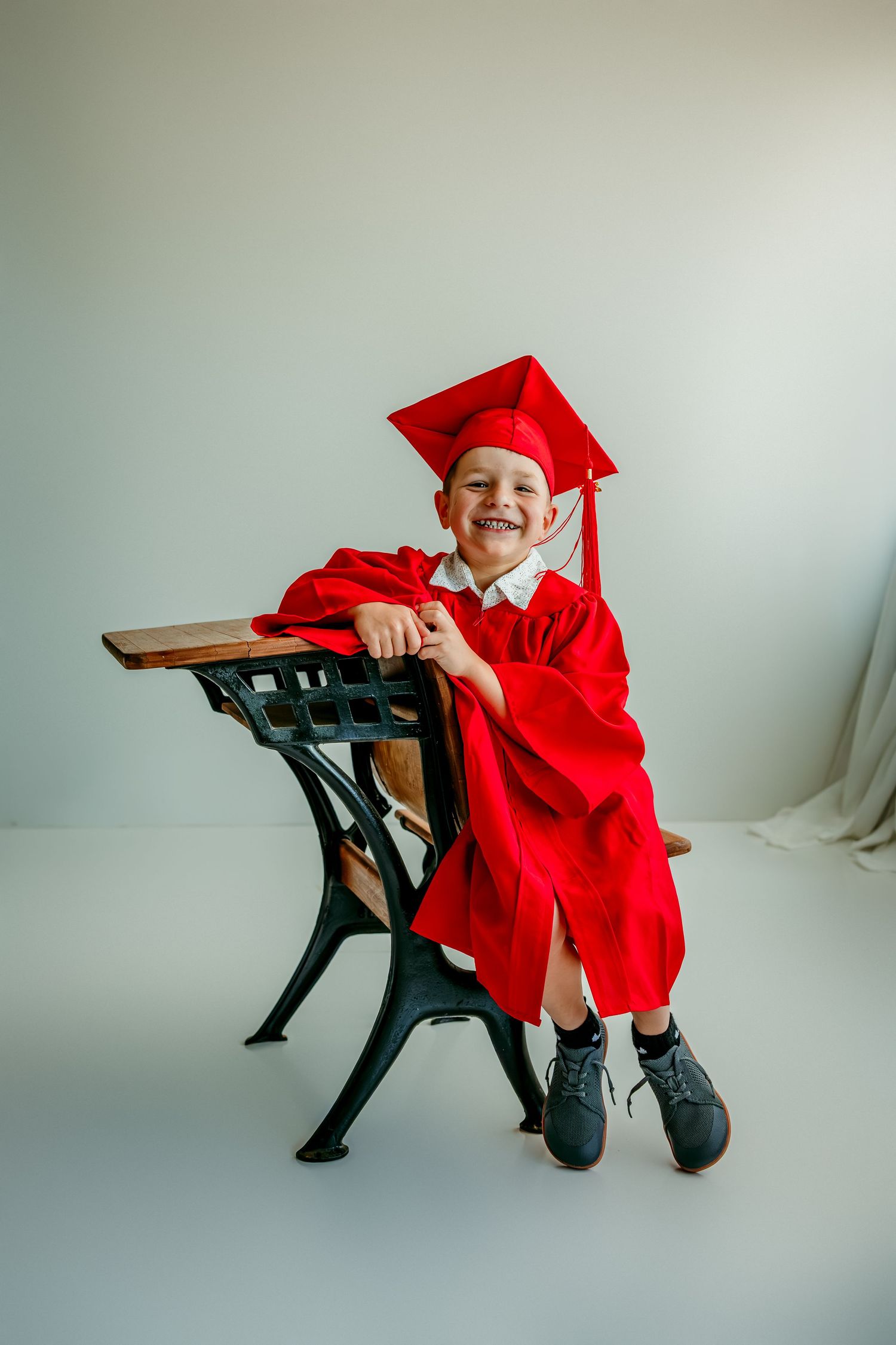 Smiling child in red graduation cap and gown sitting at a vintage school desk in a bright studio.