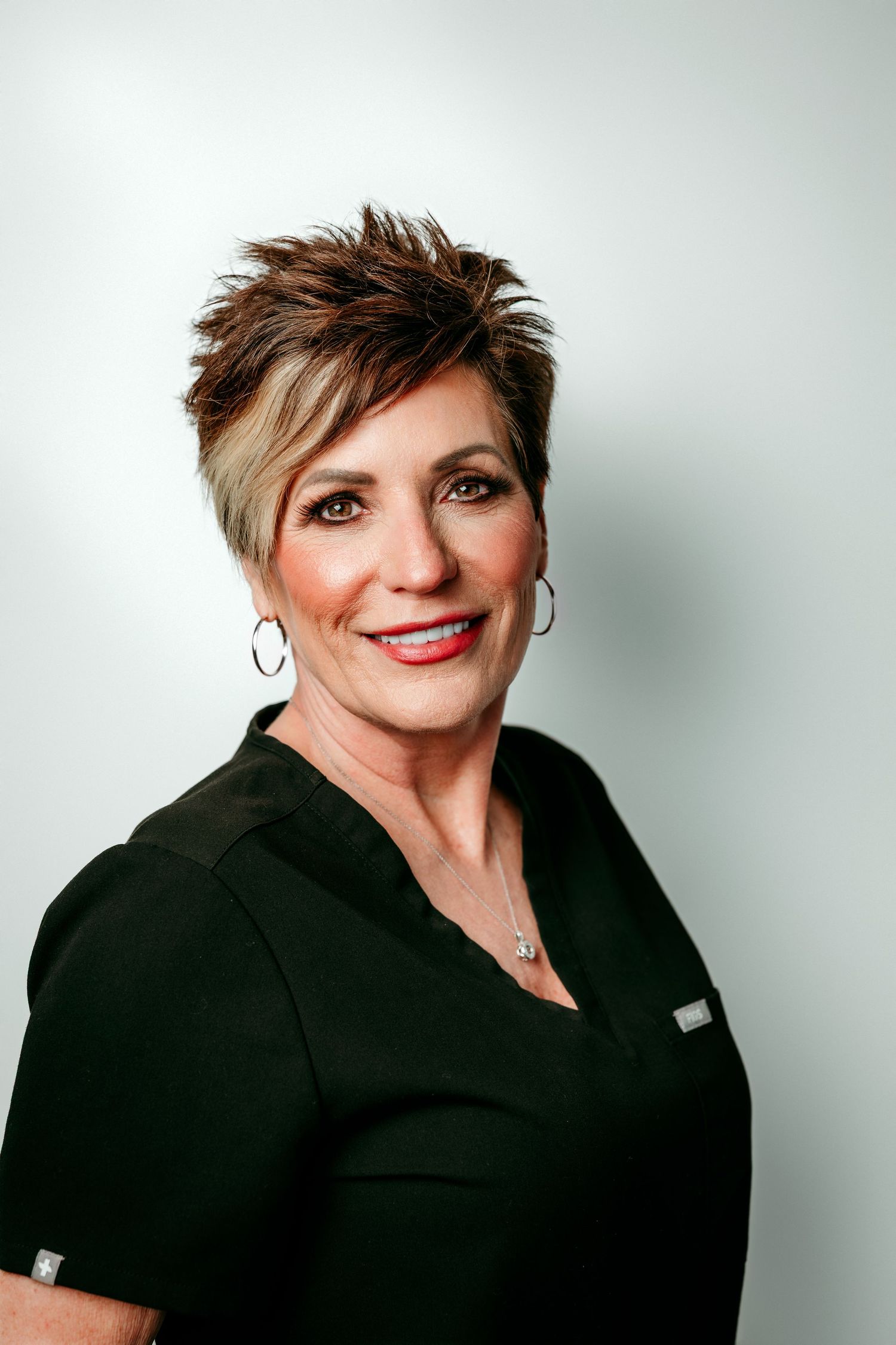 A smiling woman with short spiked hair wearing a black scrub top and hoop earrings poses for a professional headshot.