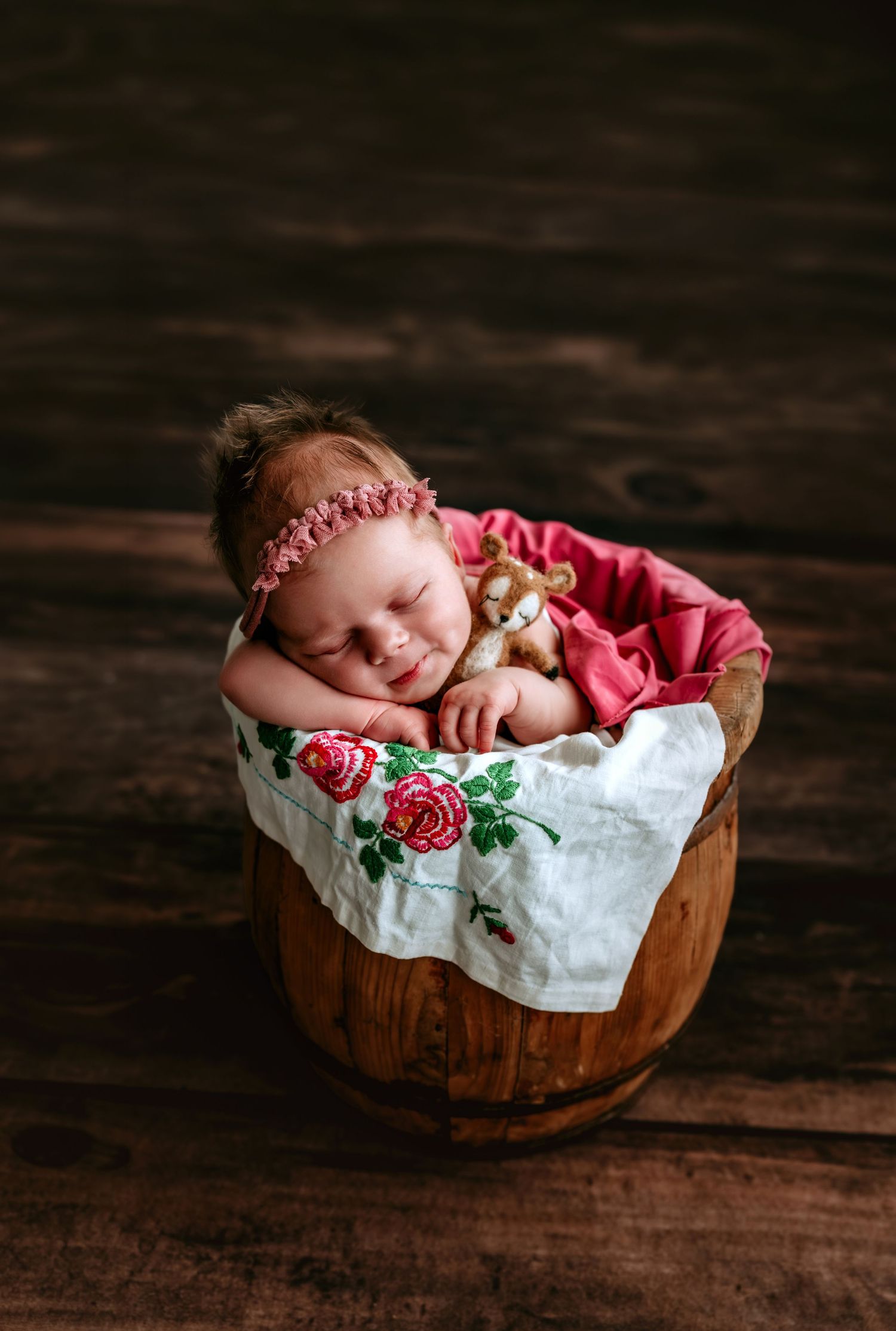 Sleeping newborn baby girl in a wooden barrel with a floral blanket, pink dress, headband, and stuffed bear prop.