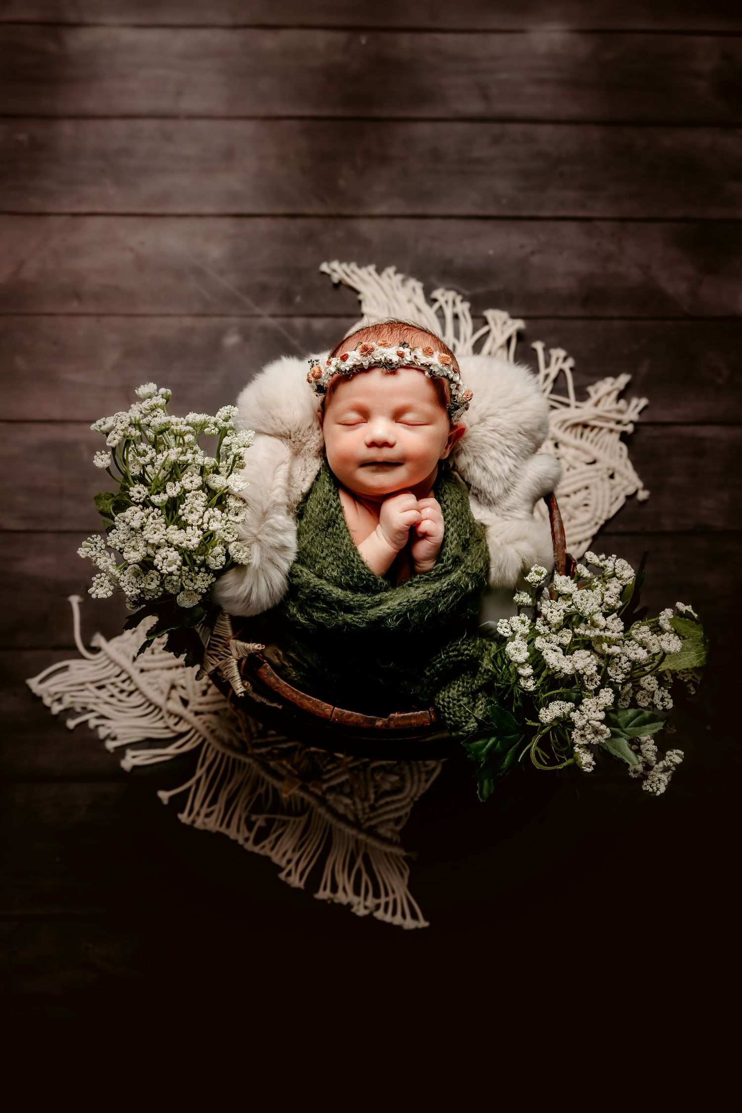 Newborn photography with green wrap and white flowers in rustic wooden basket on dark background.