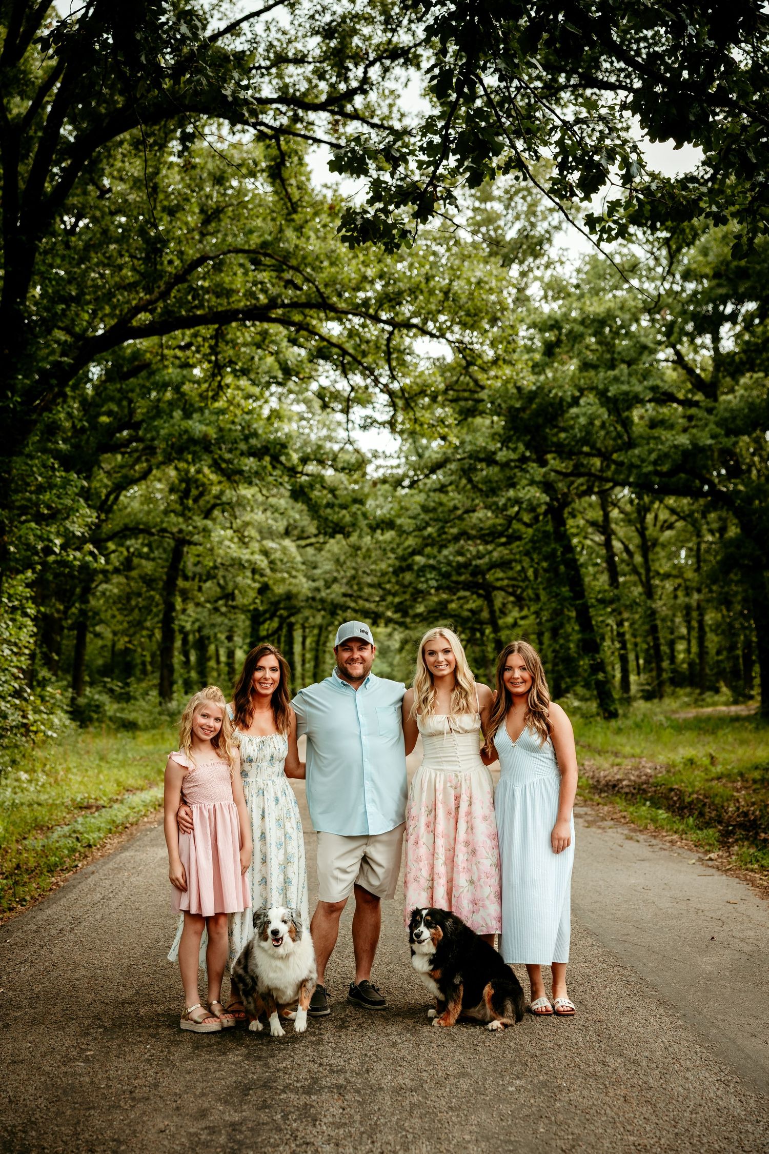 Family of five poses with two dogs on a tree-lined path, wearing pastel outfits for an outdoor portrait.