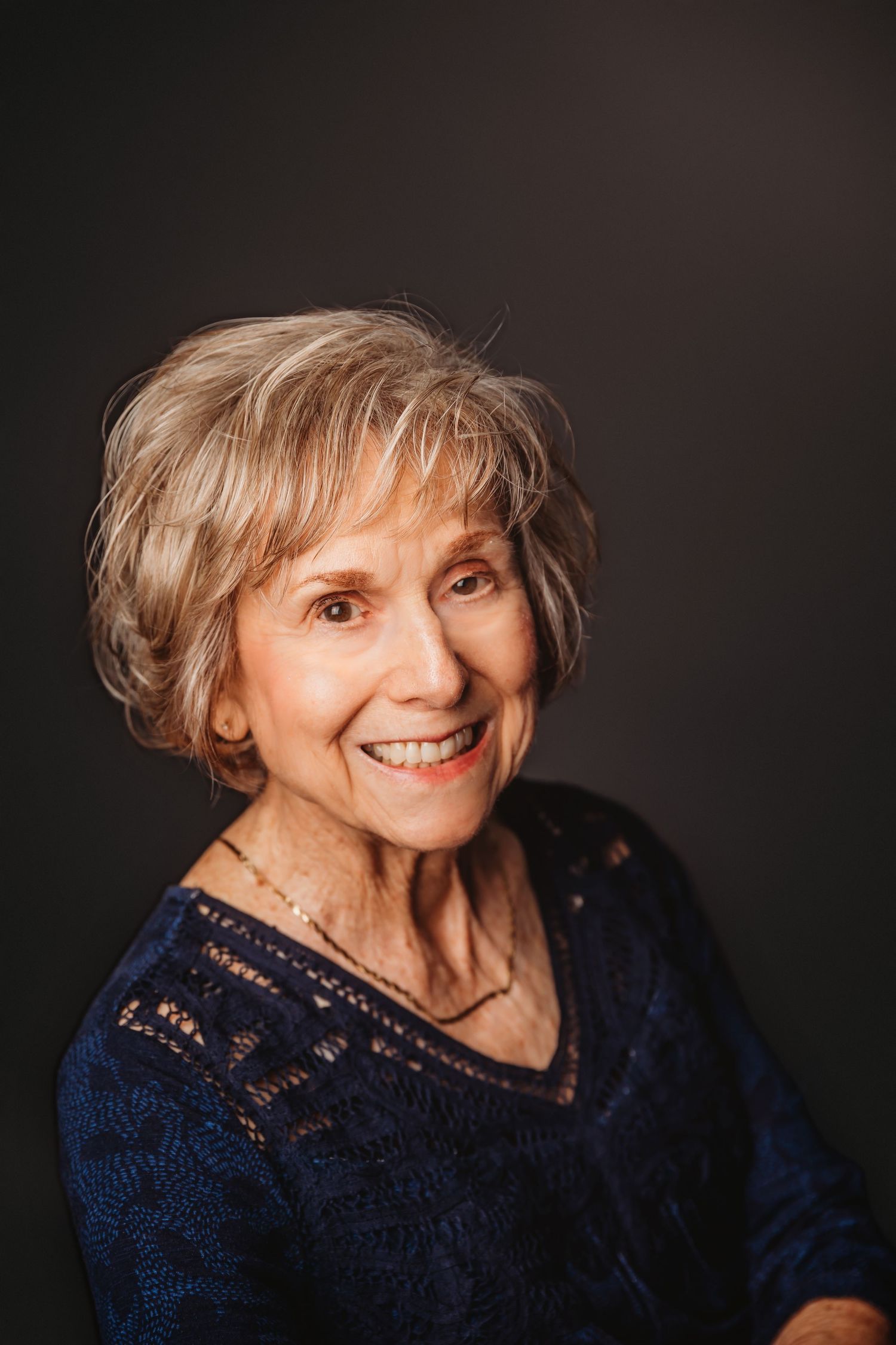 Smiling elderly woman with short gray hair wearing a blue lace top, posed against a dark studio background.