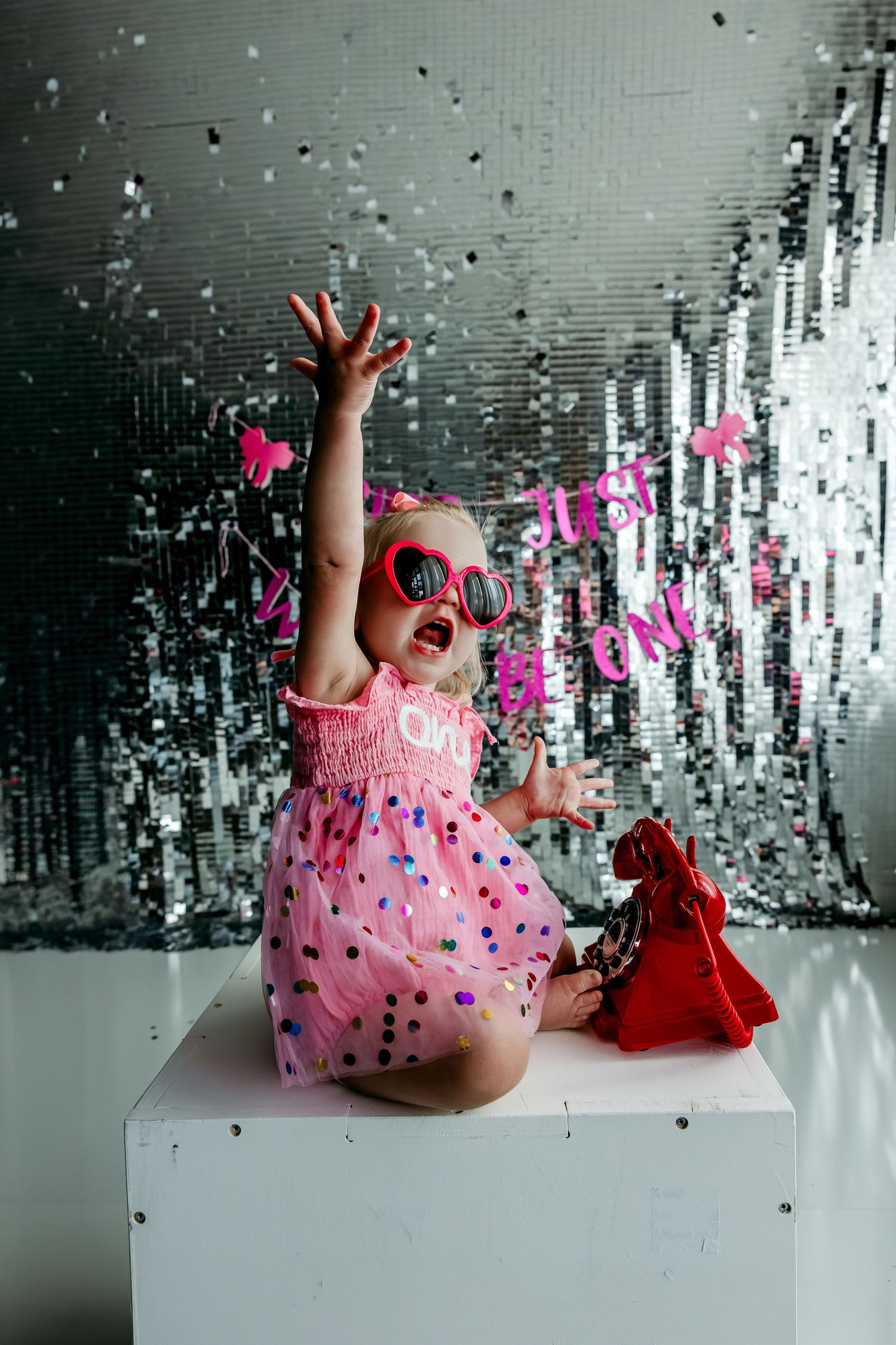 Baby girl in pink polka dot dress and heart sunglasses celebrates first birthday with confetti on sparkly backdrop.