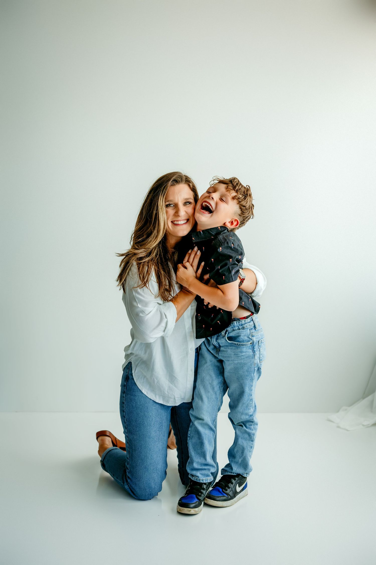 A woman kneeling on a white studio floor hugging a laughing young boy in jeans and a dark shirt.