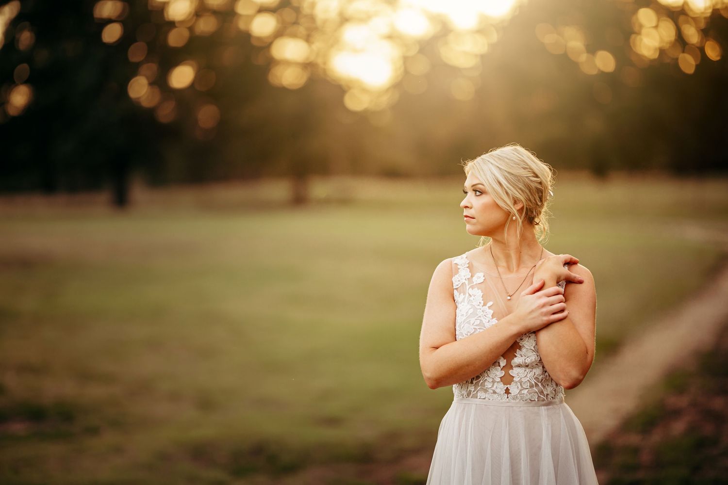 Bride in floral lace gown stands in sunlit field at golden hour, hands crossed over heart.