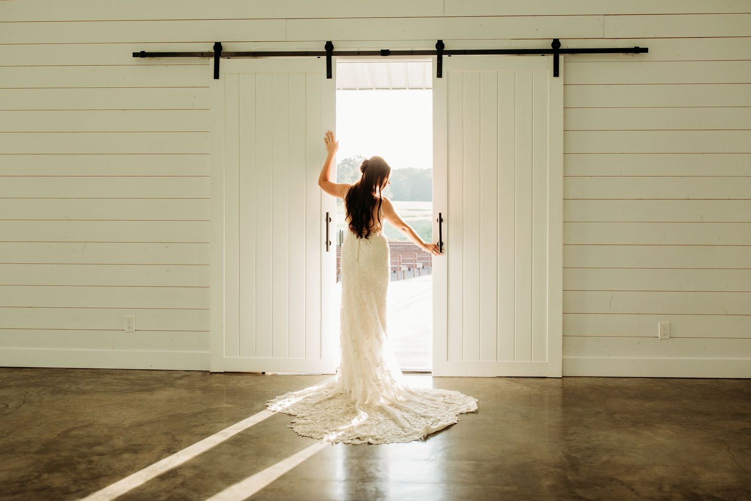 Bride in white lace gown opening barn doors to sunlight in a modern white venue with concrete floors.
