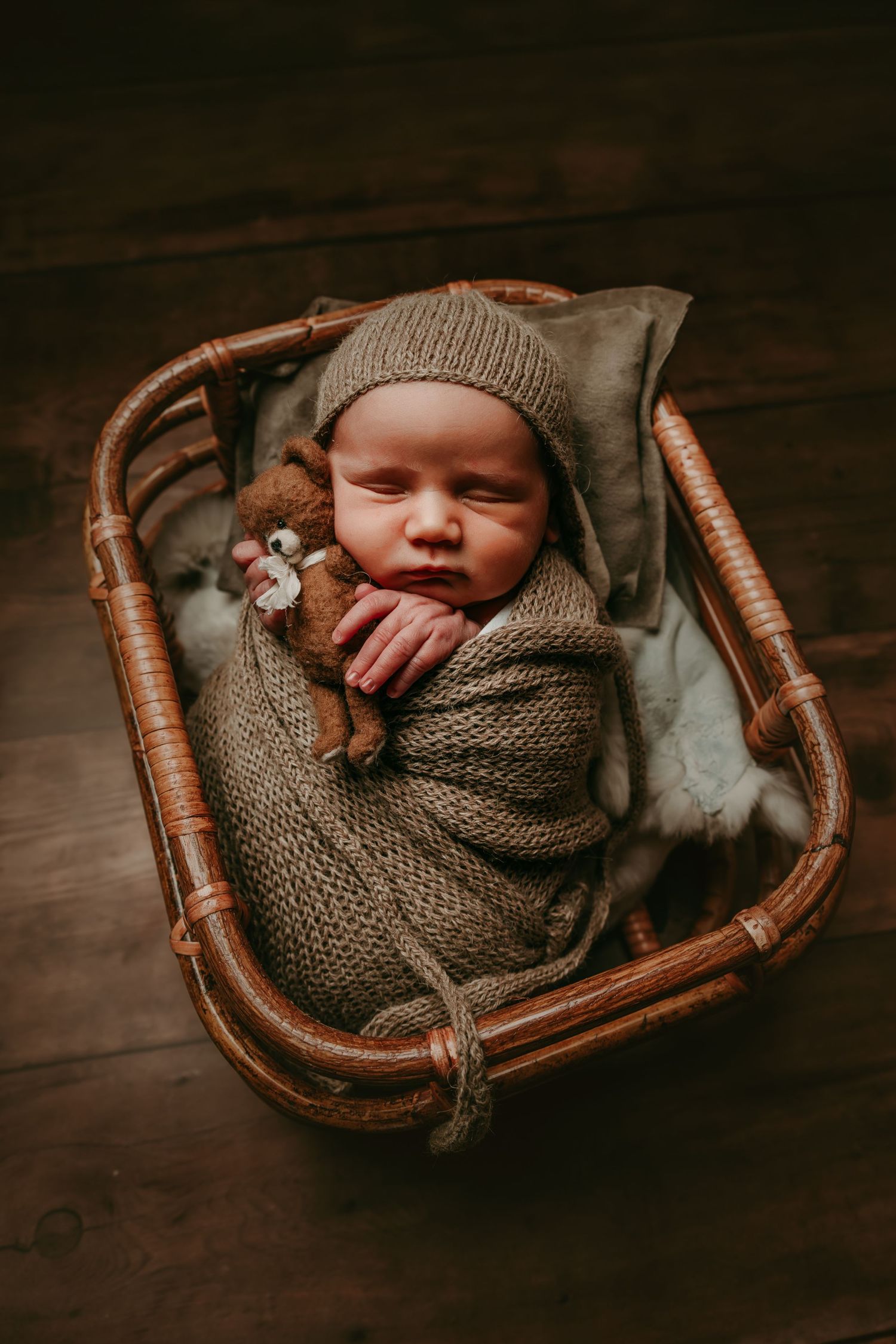 A sleeping baby wrapped in a cozy knit blanket rests peacefully in a vintage rattan basket with a small plush toy.