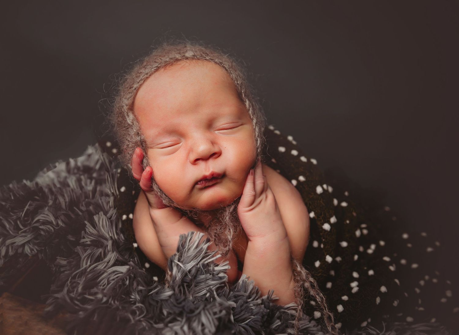 Sleeping newborn baby wrapped in soft gray fur blanket wearing a delicate knit bonnet in a professional studio portrait.
