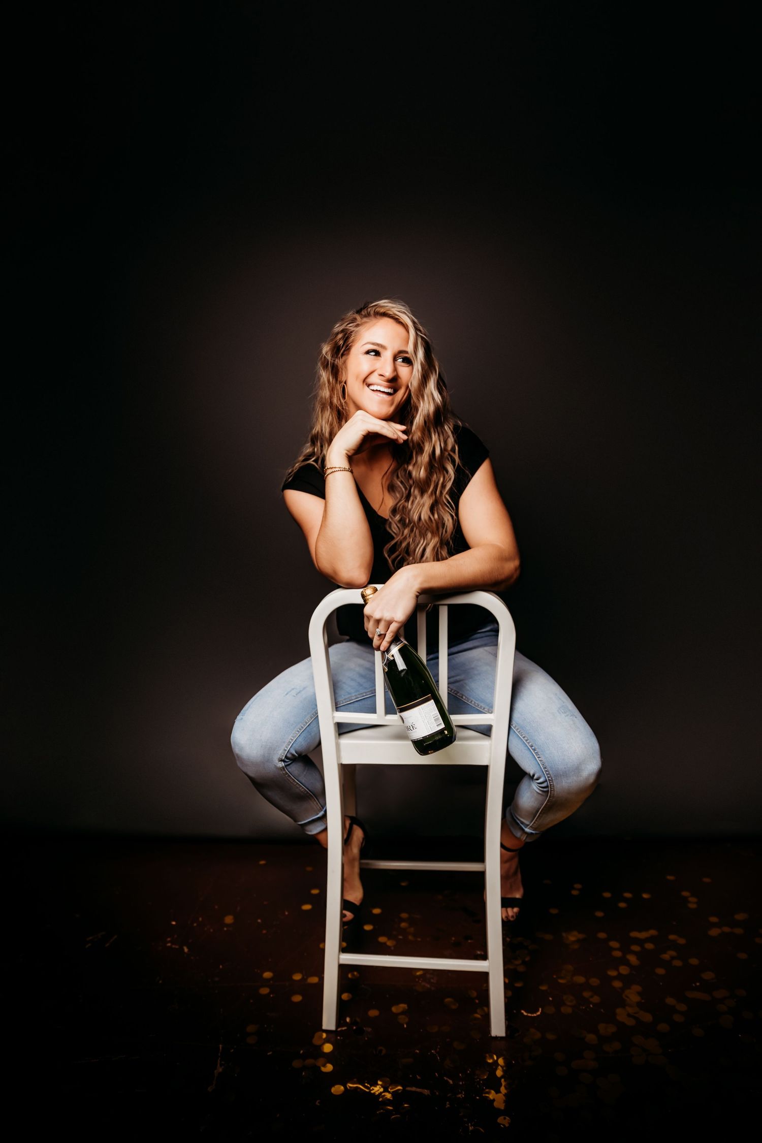 Smiling woman sitting on white chair holding wine bottle, wearing jeans and black top against dark background.