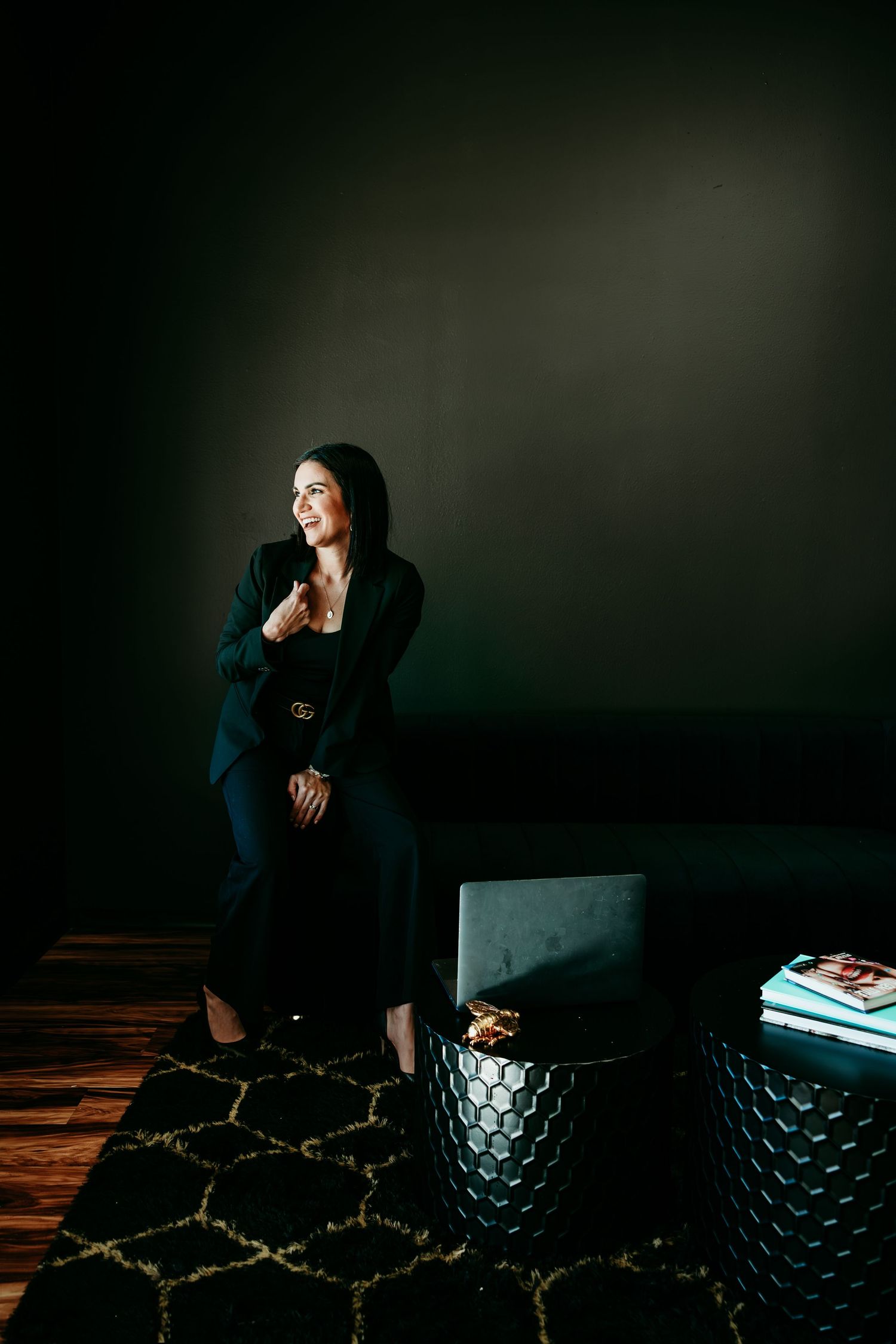 A woman in a black suit sits confidently in a dark, moody room with modern decor and a patterned rug.