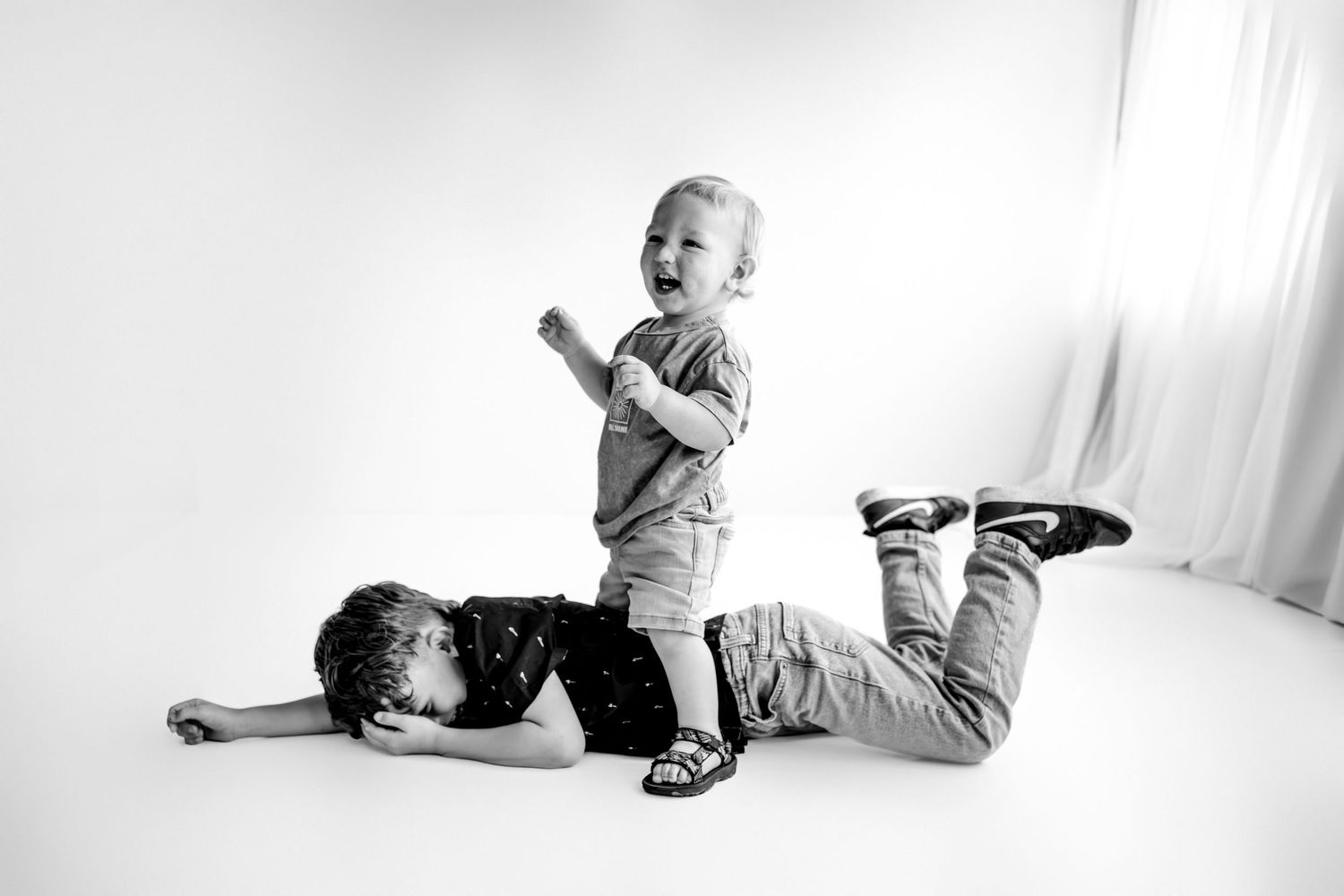 Black and white photo of two children playing, one lying on floor while a laughing toddler stands on their back.