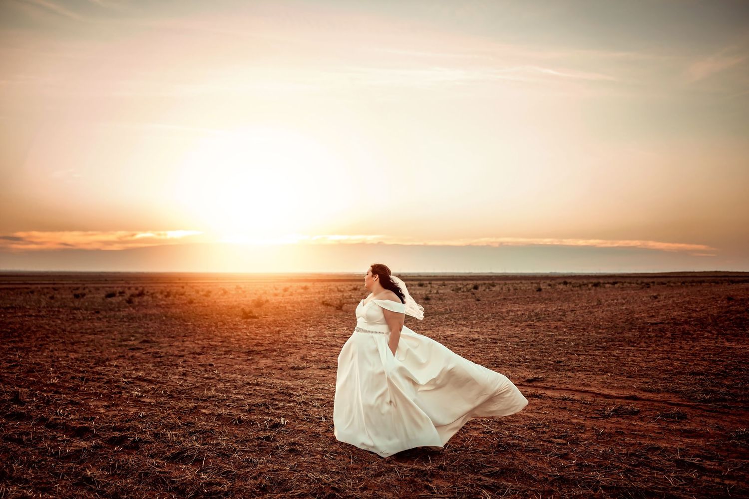 Bride in white gown twirling in open field at golden sunset, dress flowing in wind.