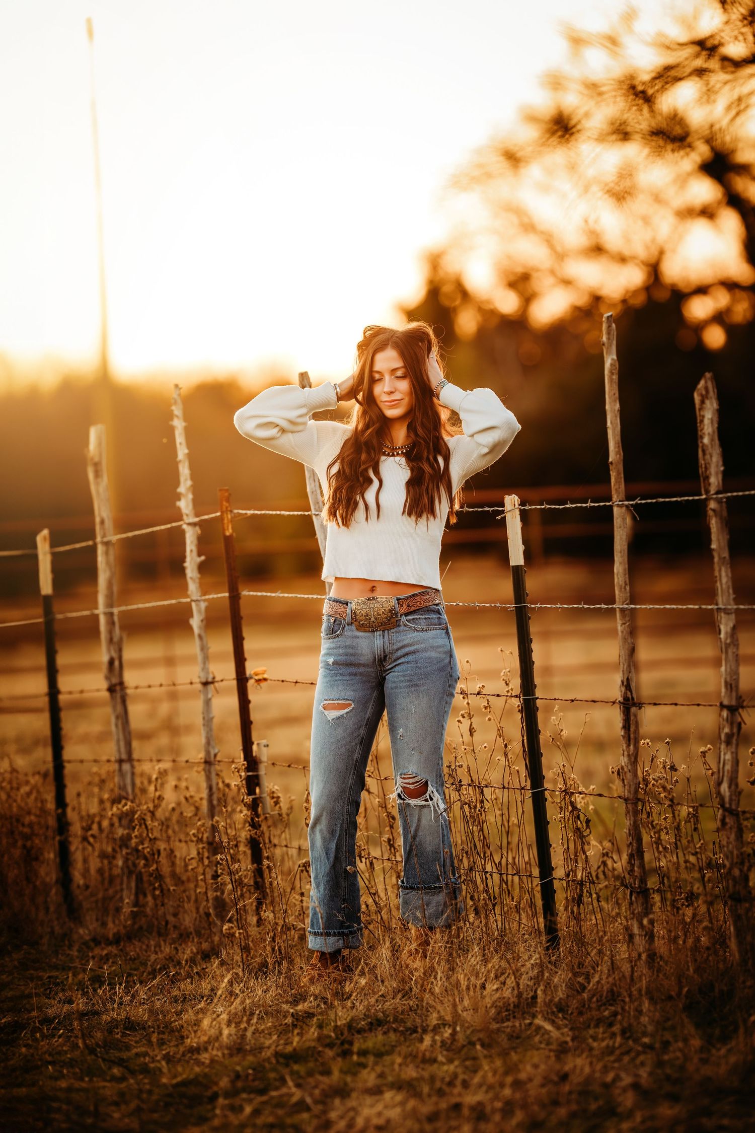 Young woman in white crop top and ripped jeans poses by rustic fence post at golden sunset.