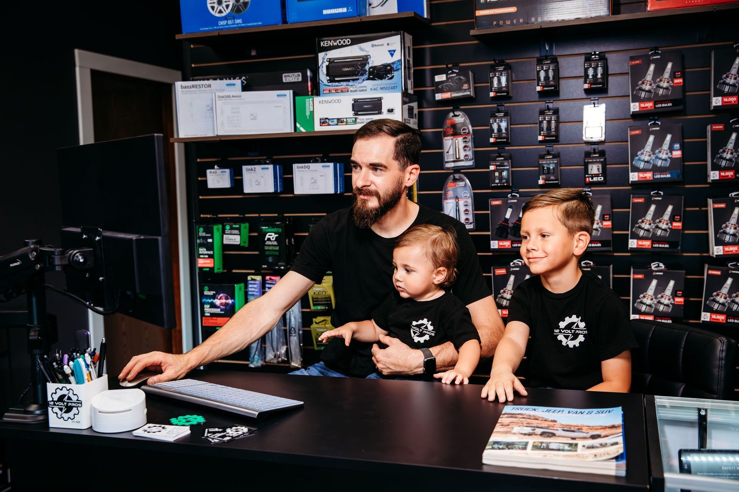 Man with two children at a retail store counter, all wearing matching black logo shirts, surrounded by automotive accessories.