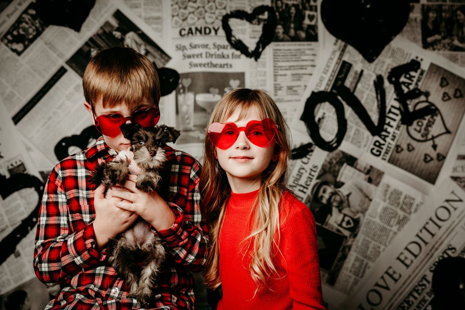 Two people in red and plaid outfits pose together against a wall covered in black and white newspaper clippings.