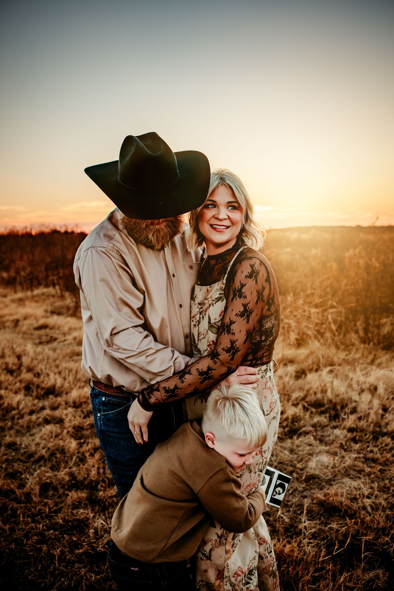 Family portrait at golden hour in a field, man in cowboy hat embracing smiling woman while child hugs them both.