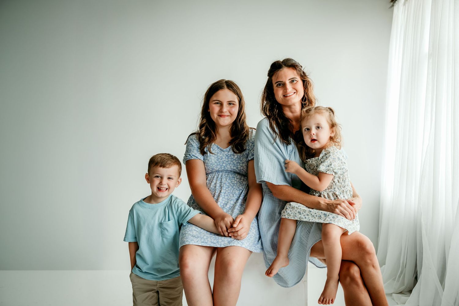 A smiling mother poses with her three young children in a bright, airy studio wearing coordinating blue and white outfits.