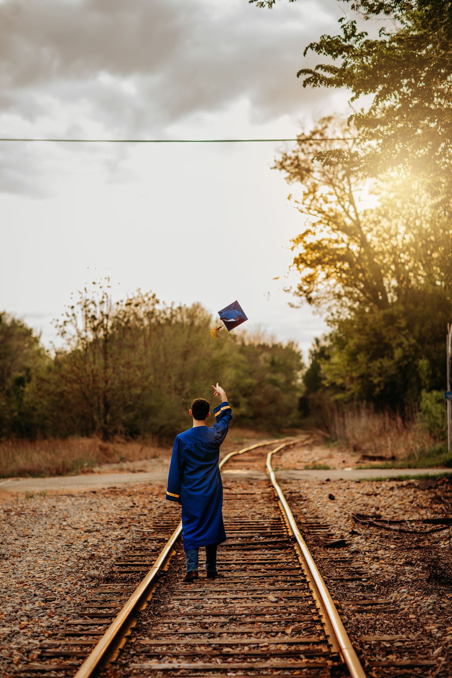 Graduate in blue gown tosses cap on rustic railroad tracks at sunset, trees lining the path.