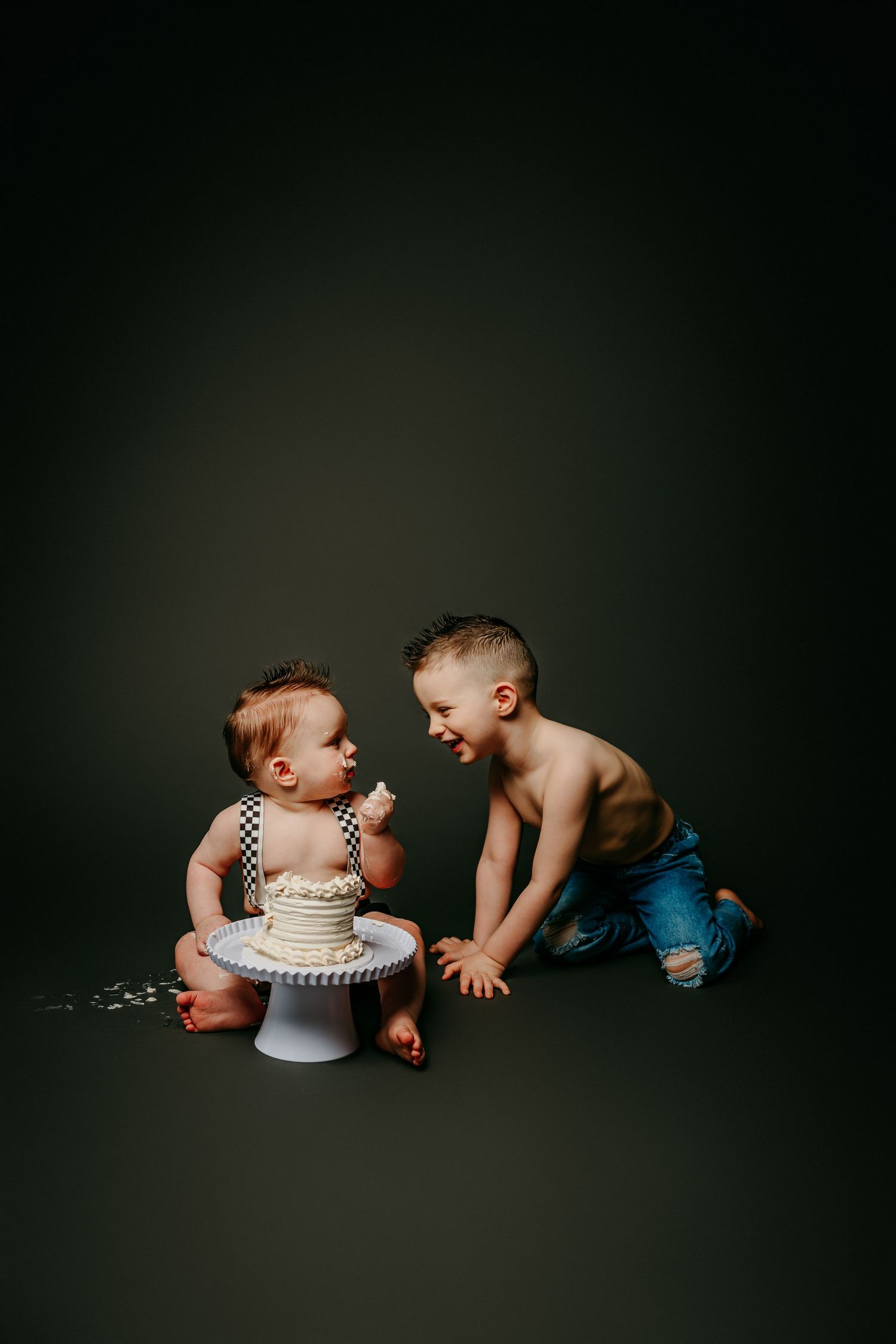 Two young boys sharing a cake smash birthday celebration, with a white frosted cake on a pedestal against a dark background.