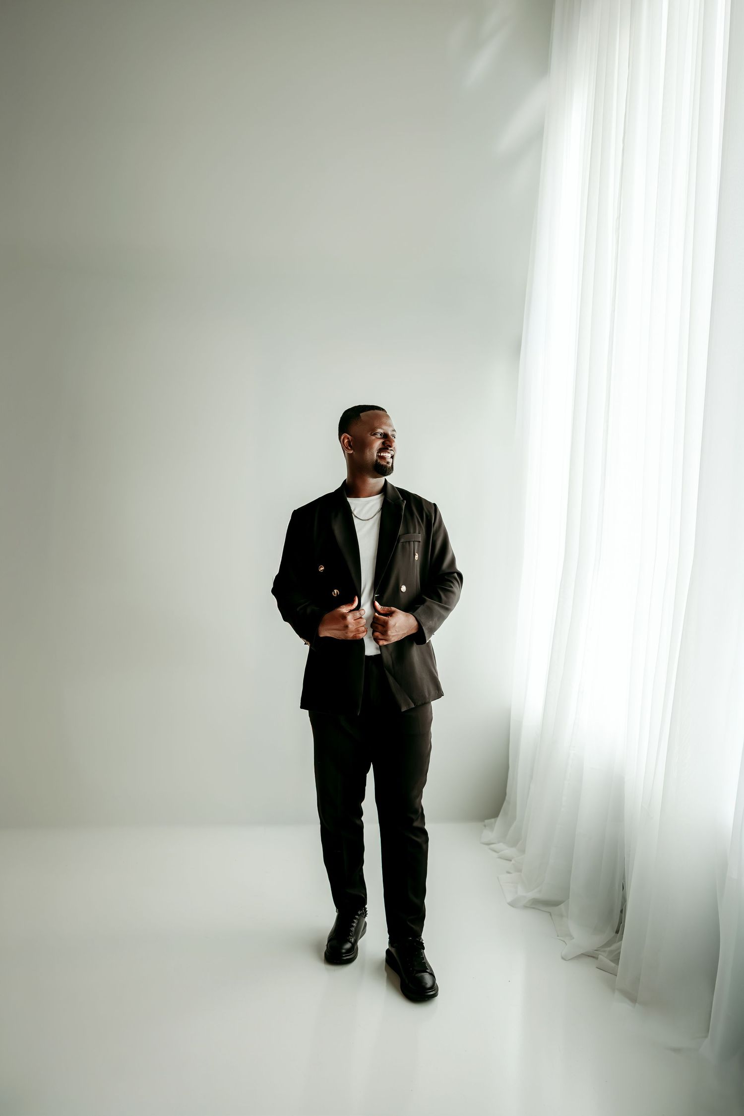 A well-dressed Black man in a dark blazer and trousers stands confidently in a bright white studio near sheer curtains.