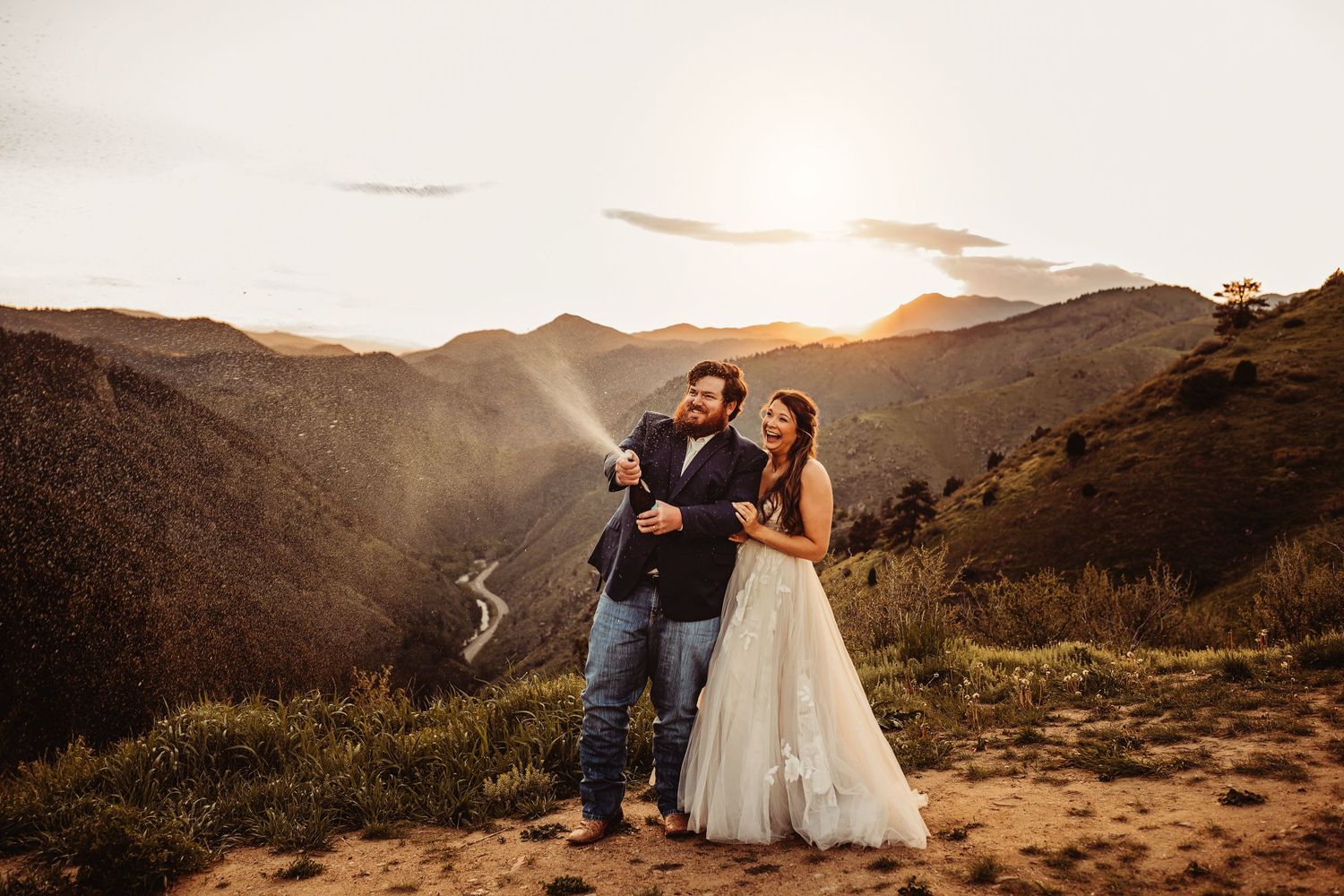 Couple celebrating wedding at sunset on mountain overlook, bride in white gown, groom in suit, golden light over canyon.