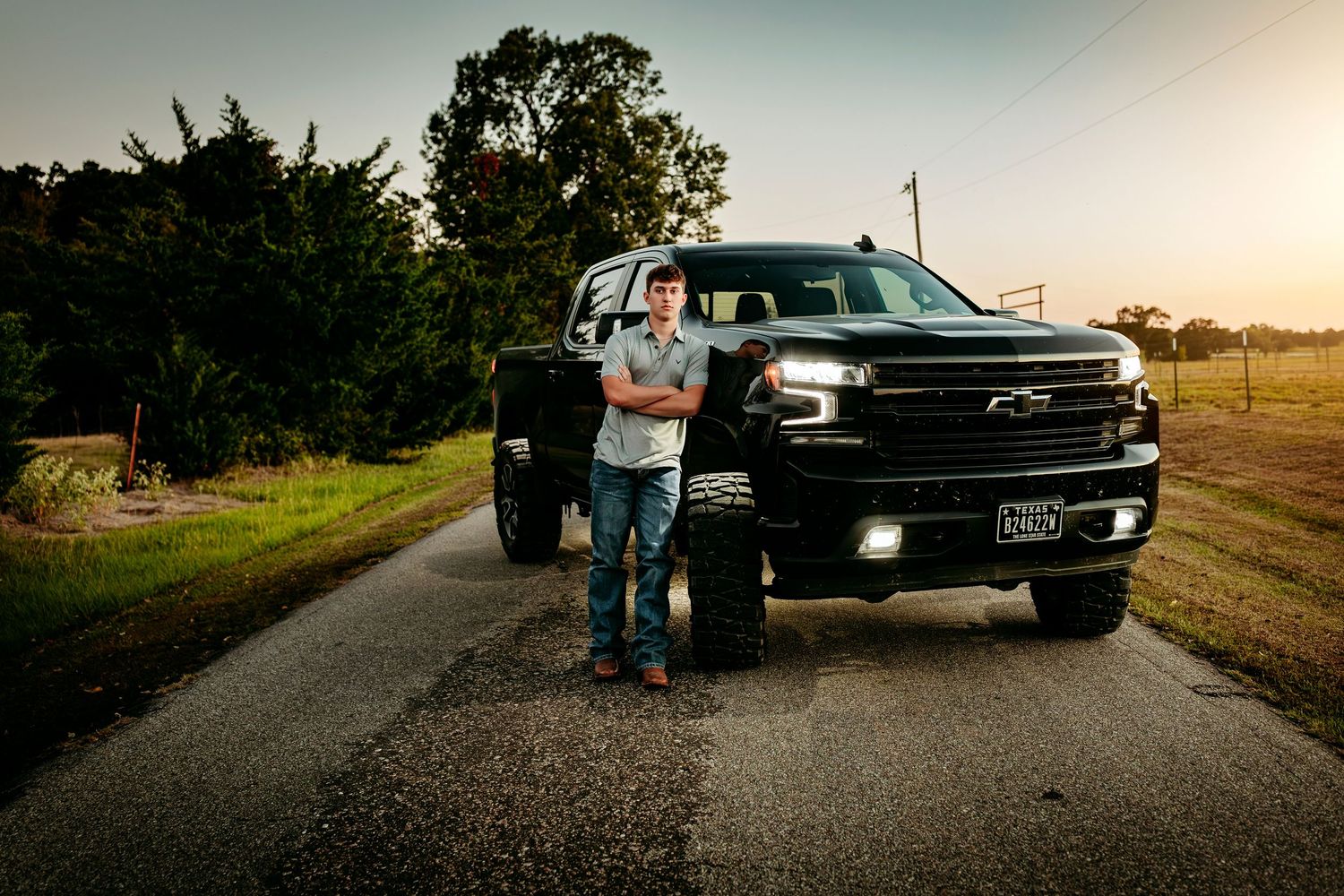 Young man standing with arms crossed in front of a black lifted Chevrolet Silverado truck on a rural road at sunset.