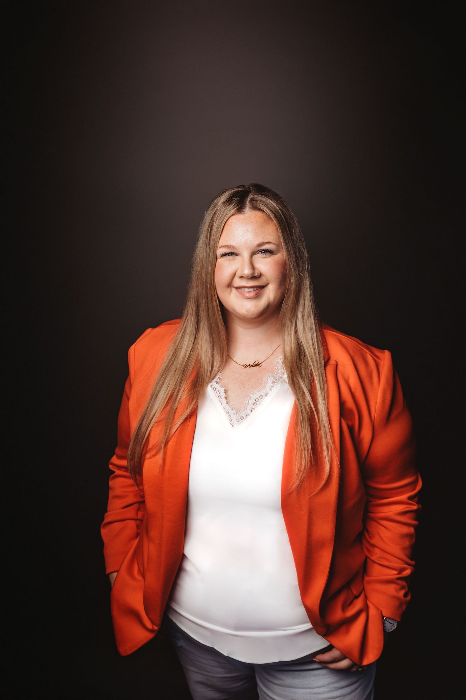 Woman in orange blazer and white top smiling confidently against dark background in professional portrait photo.