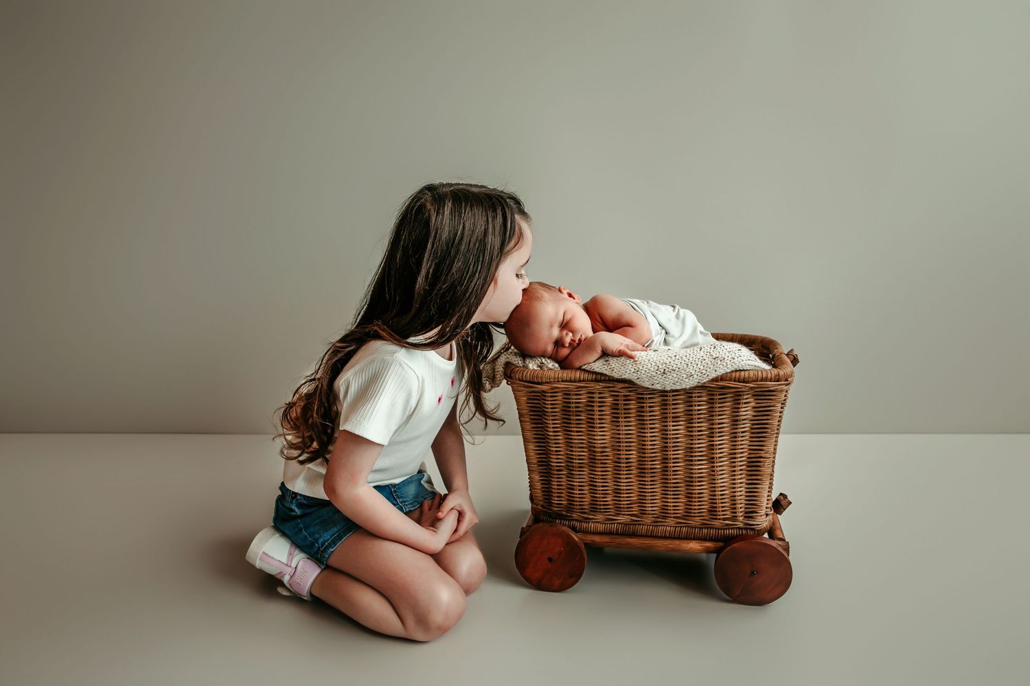 Young girl sitting on floor kissing sleeping newborn baby in rustic wicker wagon, tender sibling moment.