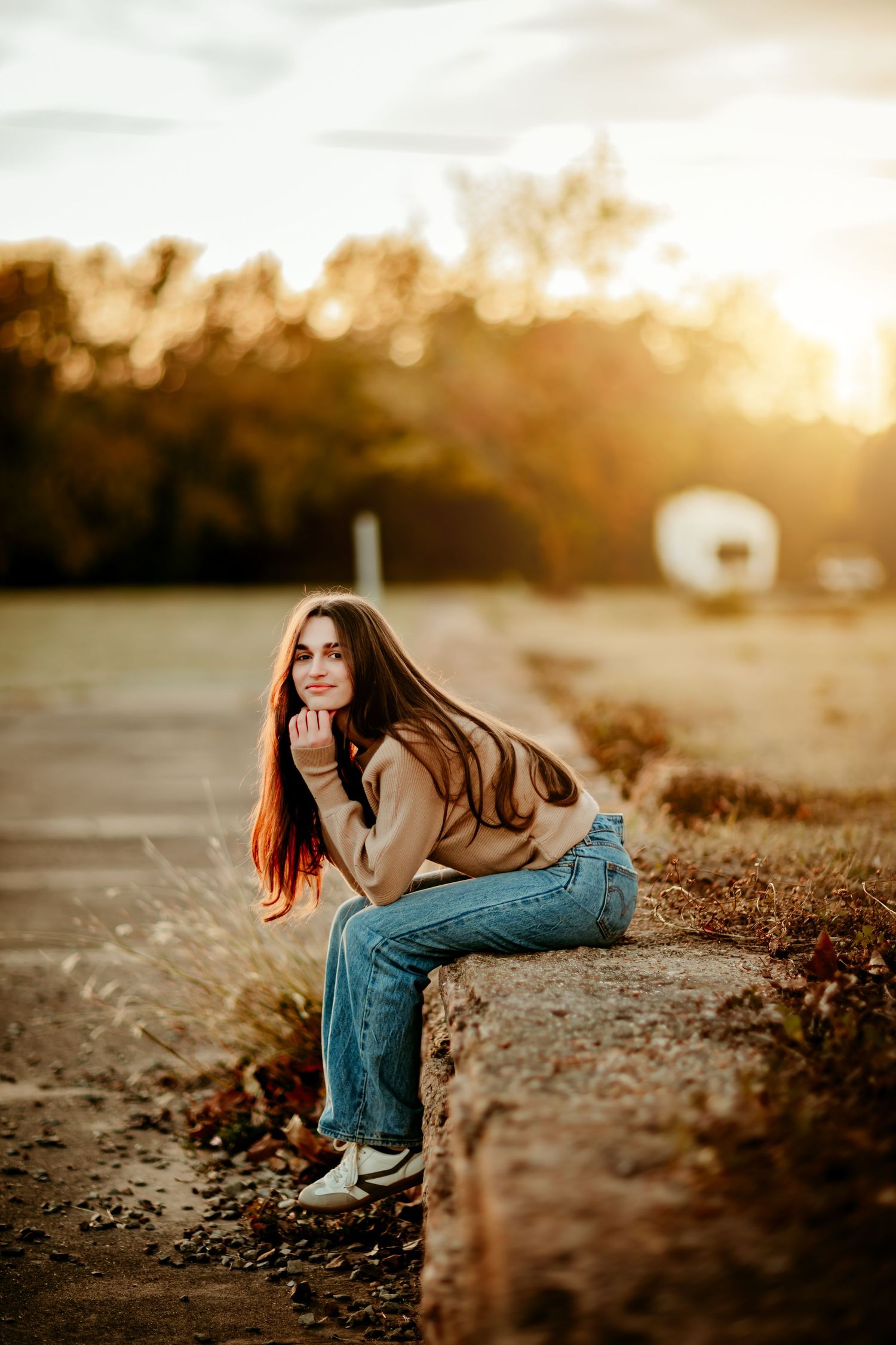 Young woman with long red hair sits on a stone ledge in jeans and tan sweater, posing at golden hour sunset.
